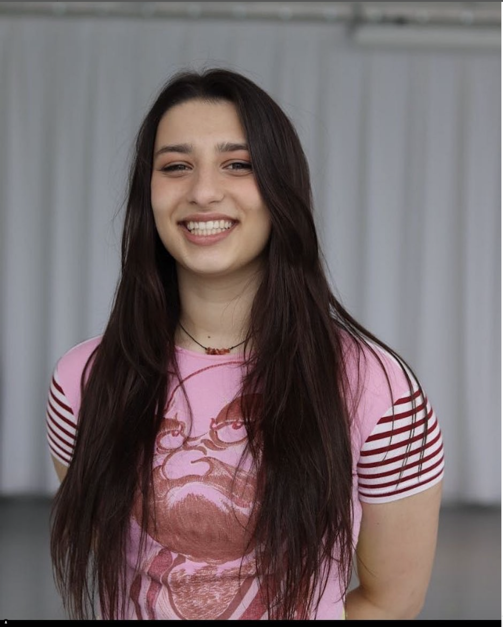 A portrait of dancer Isabella Caspariello, a white woman with long dark brown hair hanging down over shoulders. She wears a pink t-shirt, with arms behind her back and smiles broadly at us. 