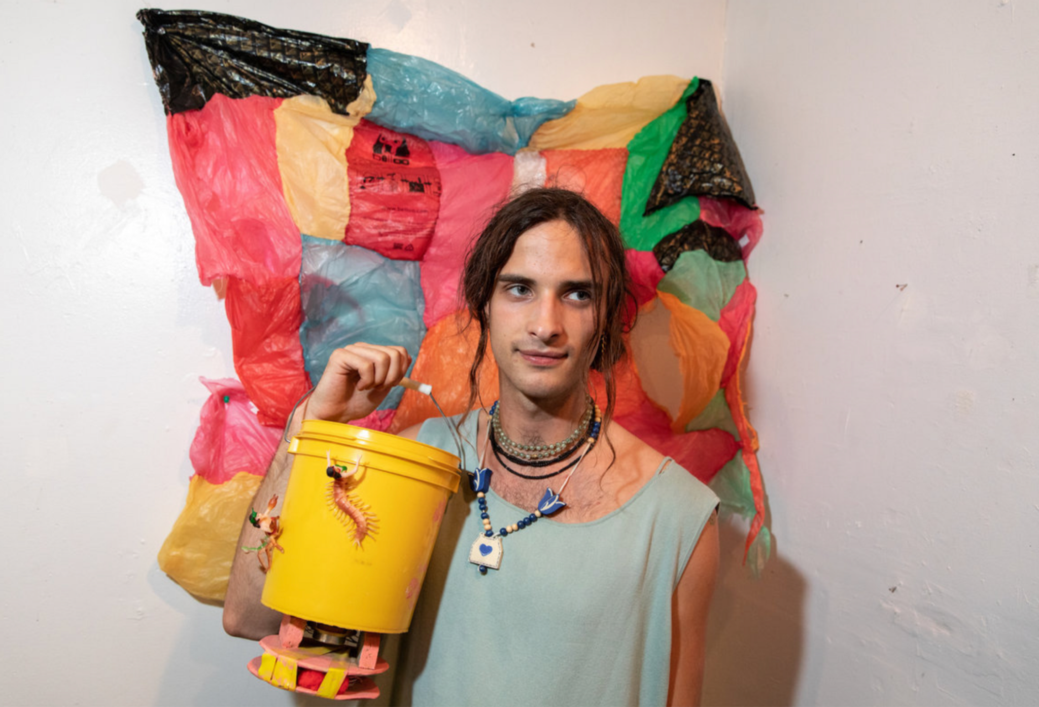 A person with long hair pulled back with strands hanging on either side of their face holds up a yellow bucket at chest level. Behind them is an artwork that looks like a multicolored patchwork quilt made of plastic bags.