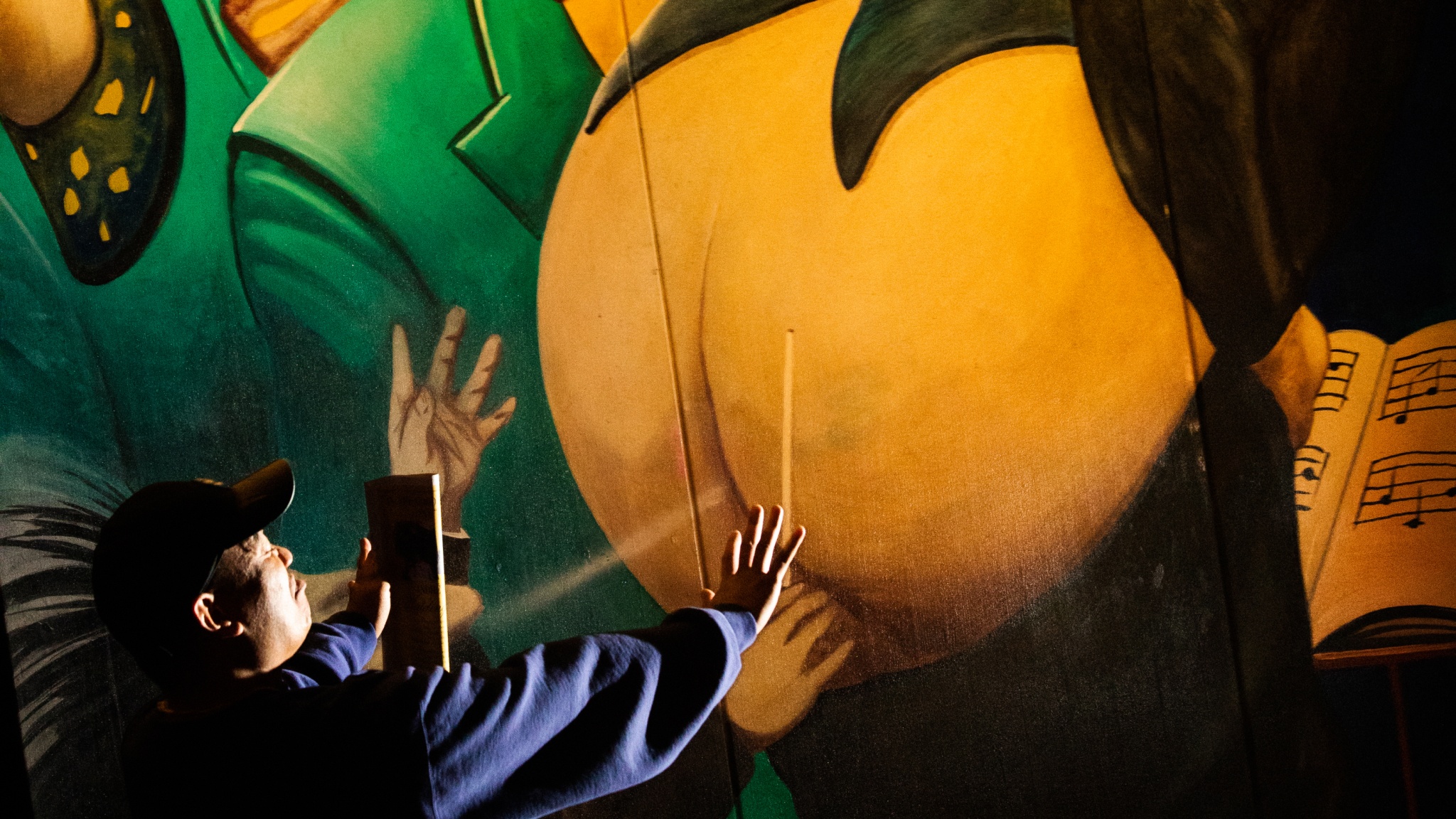 A close up of a visitor to Luna Luna interacting with a mural by Manfed Deix. In the cartoon mural image, a conductor lowers his pants to moon onlookers. The visitor holds one hand up to the mural while taking a photo with the phone in his other hand. 