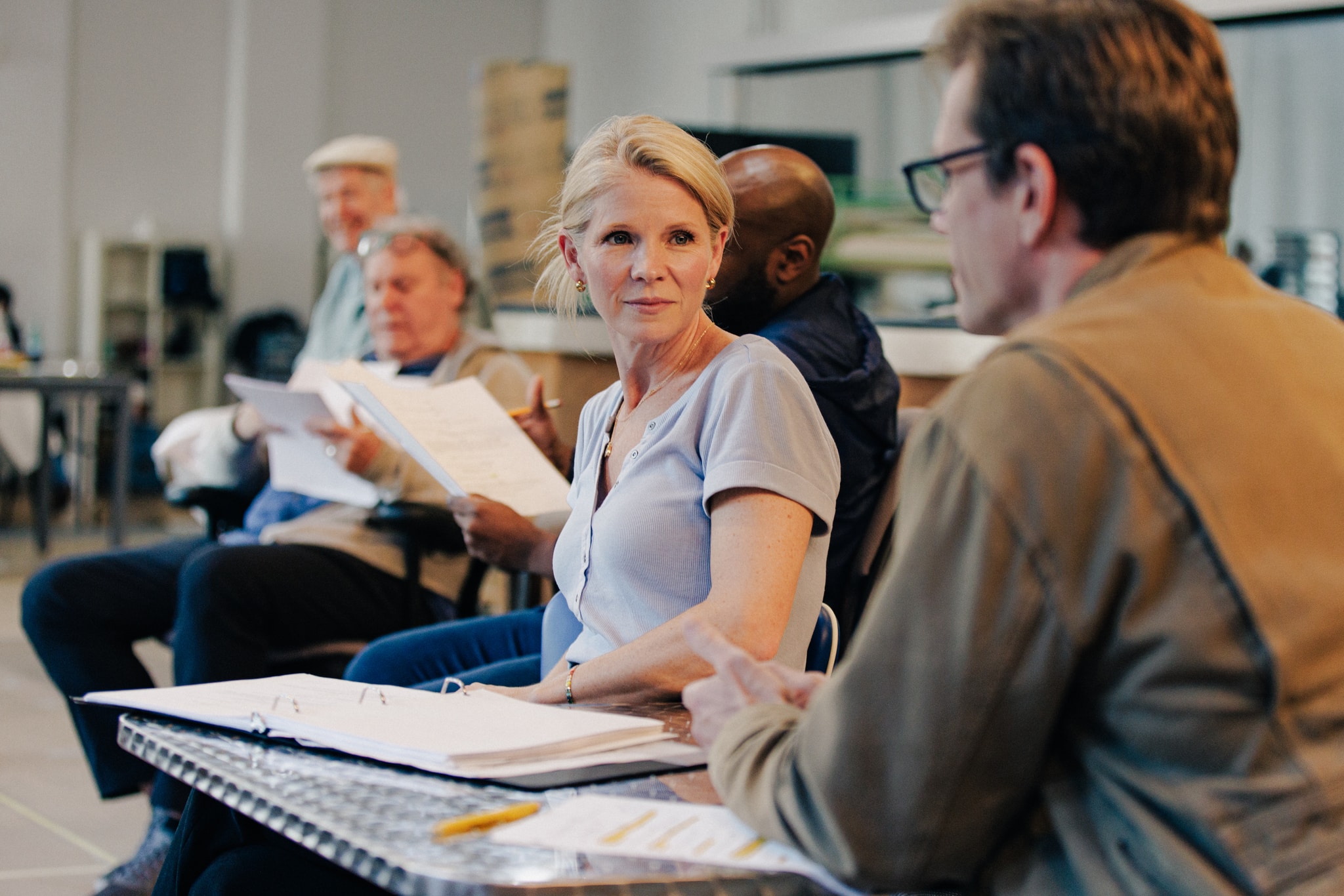 A photo of actor Kelli O'Hara turning her head to look toward us over her shoulder as she sits in a chair. In front of and behind her are lined up co-members of the cast also seated. Kelli has a vulnerable look on her face.