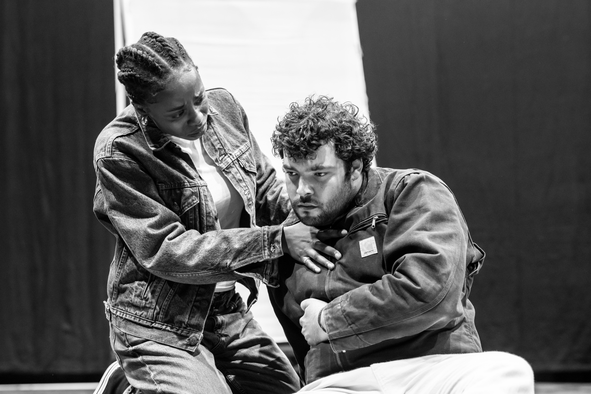 Actors Deborah Alli and Dylan Corbett-Bader rehearse a scene. Corbett-Bader is slumped on the ground as if injured and Alli kneels beside him, supporting his right shoulder. 