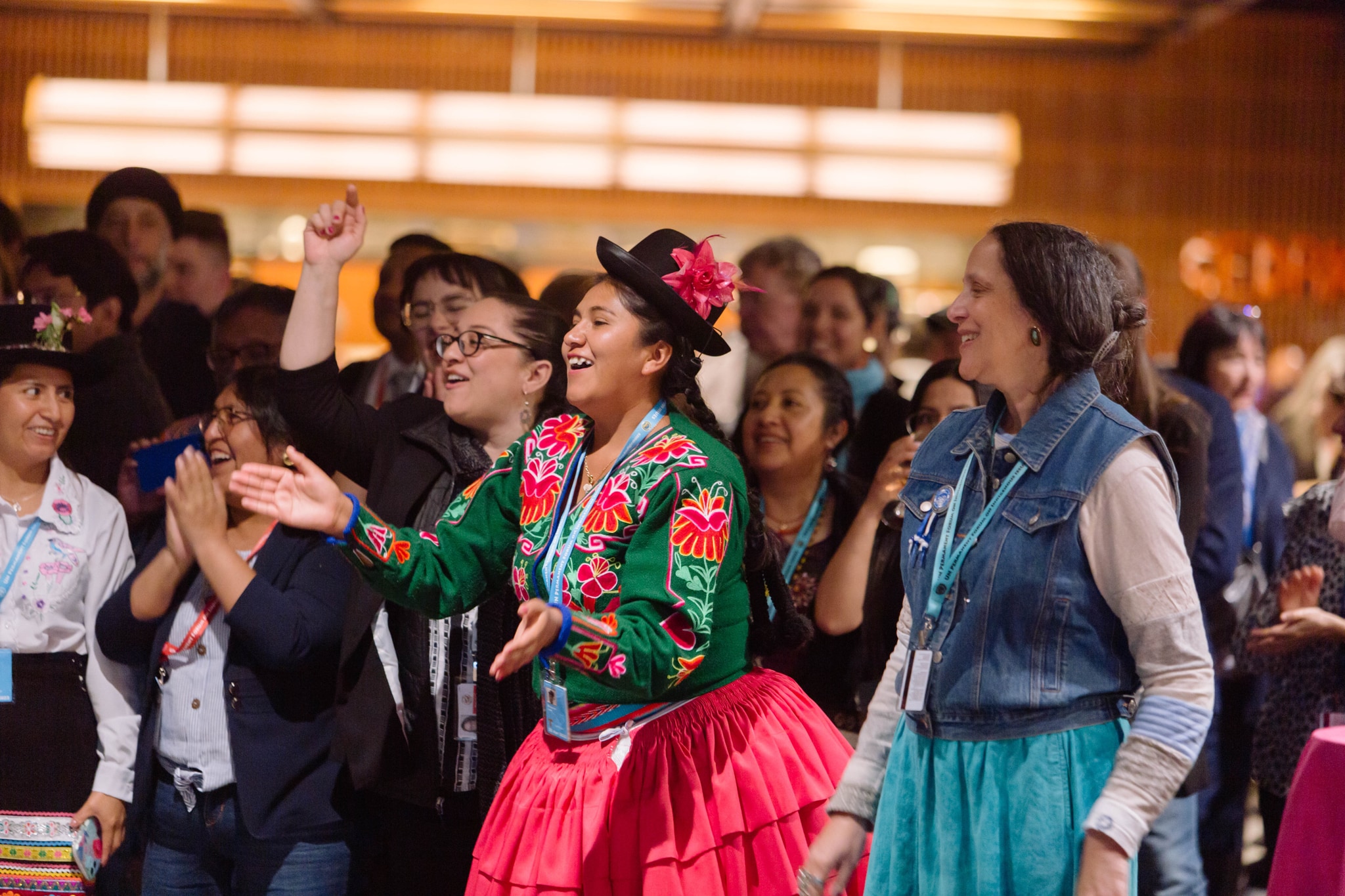 Attendees of United Nations' Permanent Forum on Indigenous Issues gathering stand in a warmly lit lobby, dancing and laughing. At center, a woman in a bright pink skirt and a green and pink jacket smiles and claps her hands.