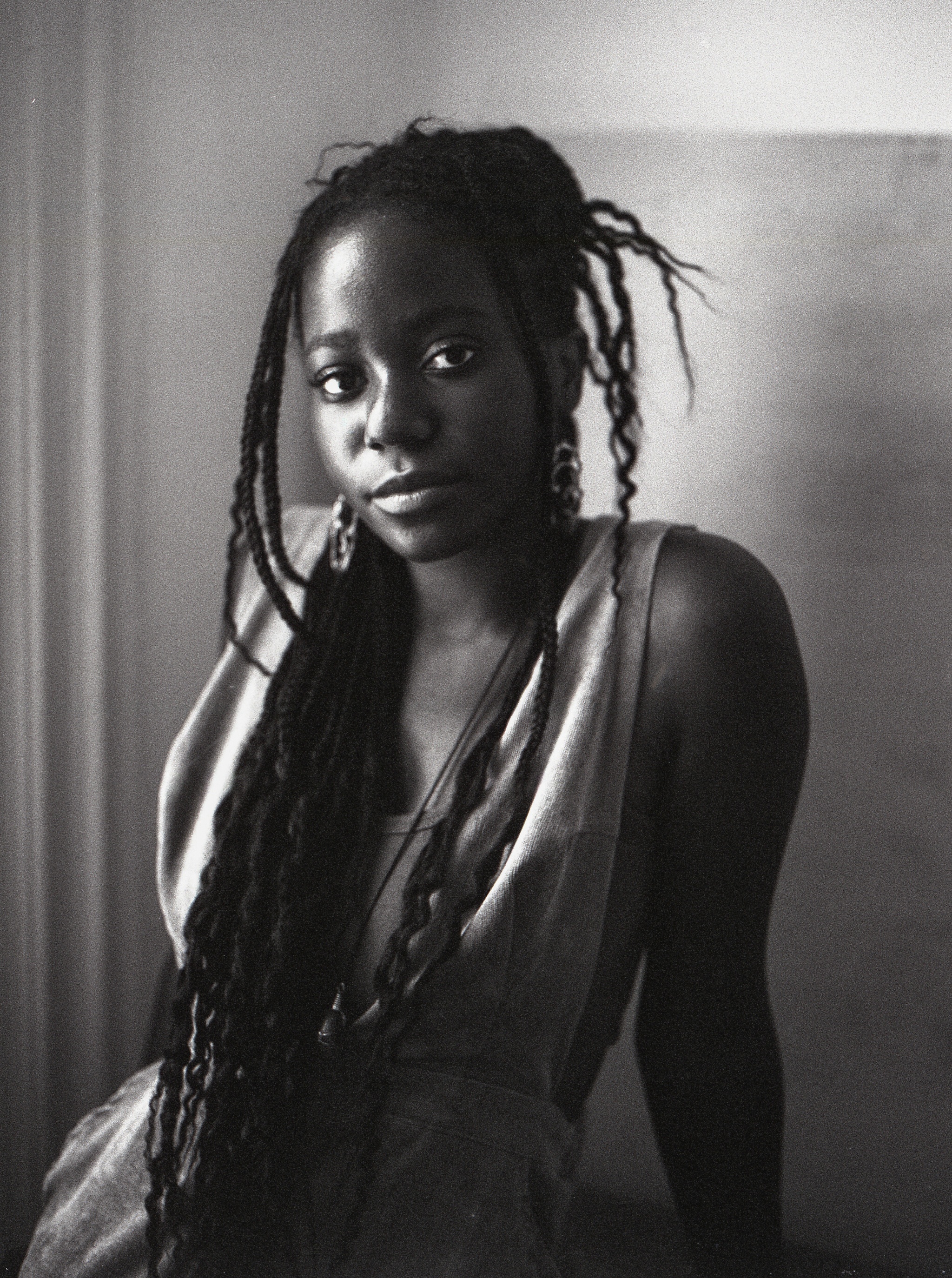 A black-and-white portrait of Haitian Canadian artist Laurena Finéus. Laurena sits with one side of her face lit by a light source out of the frame. She leans back casually, supporting herself with her arms outstretched behind her. She has long locs that fall to her waist and wears a loose-fitting dress. She looks directly at us with a pleasant look on her face. 