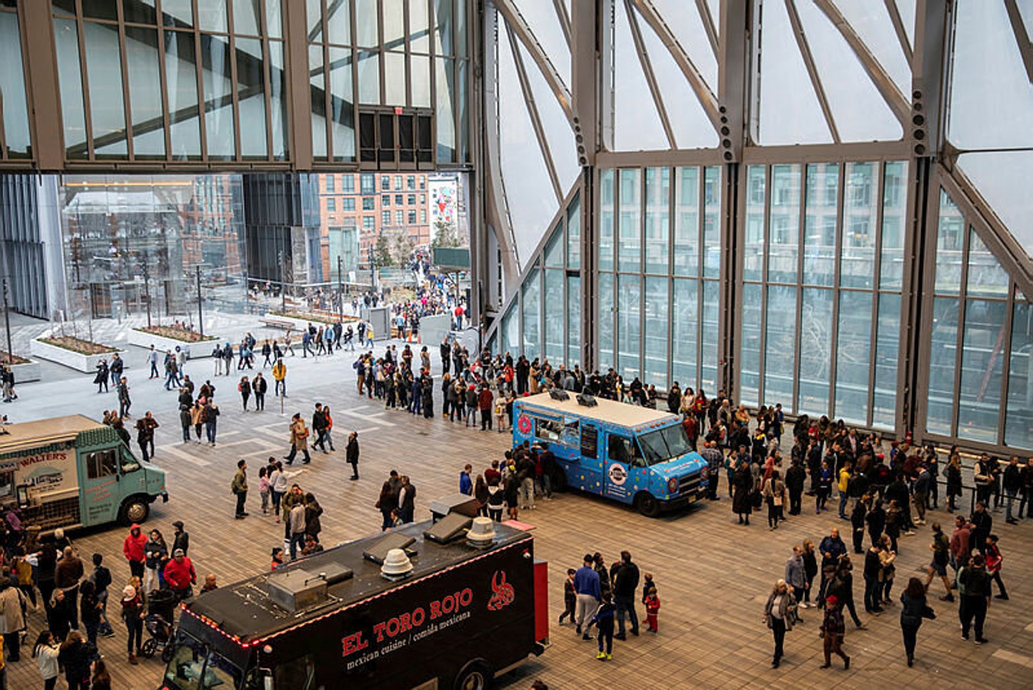In a partially enclosed plaza, crowds line up for three food trucks parked in the space. 