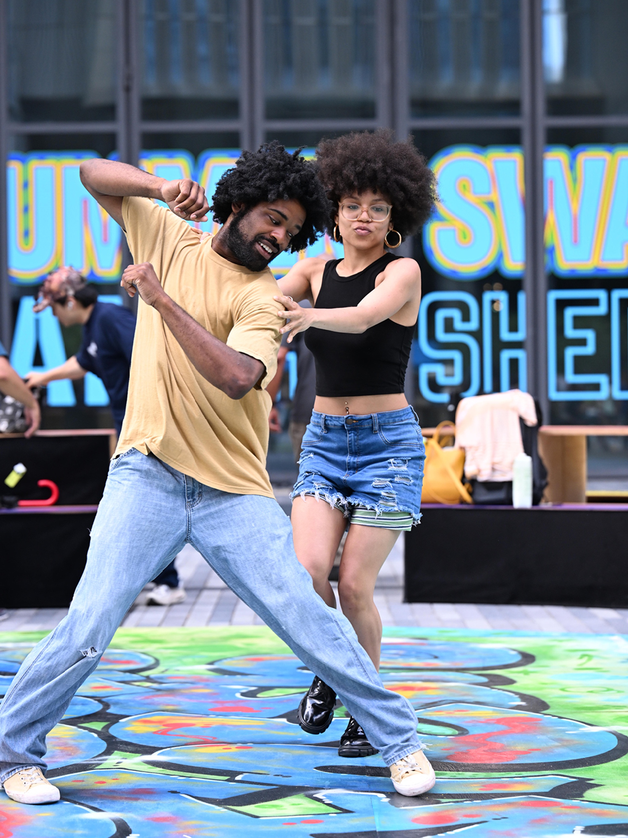 Two Black dancers, a man and a woman with Afros, perform on an outdoor plaza on a painted surface. The man, in jeans and a t-shirt, spreads his legs and holds his arms up at an angle while the woman, in a tank top and jean skirt, reaches out to his shoulder.