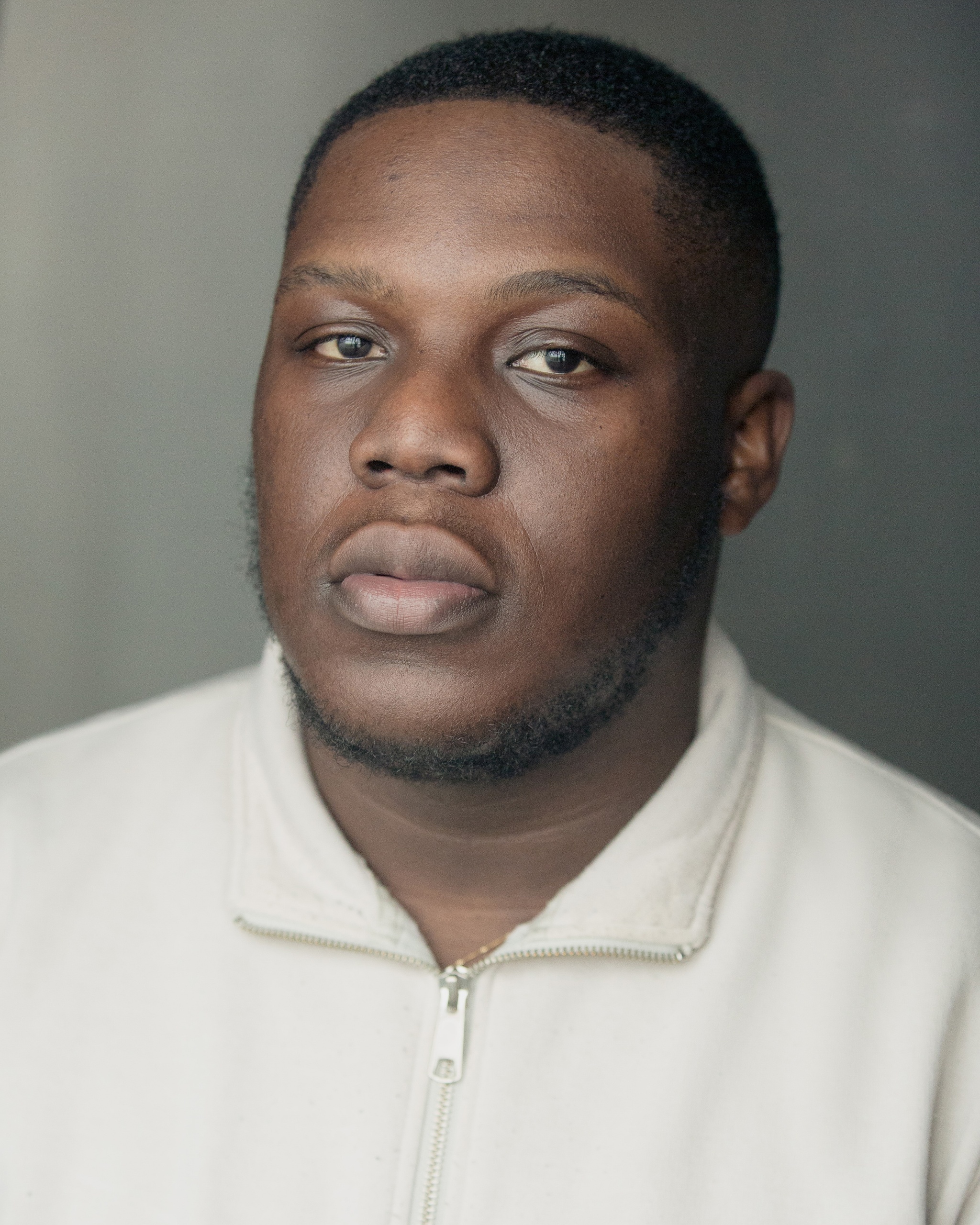 A headshot of actor Raymond Anum, who looks directly at us. Raymond has dark brown skin and wears a zip up sweater zipped to the base of his neck