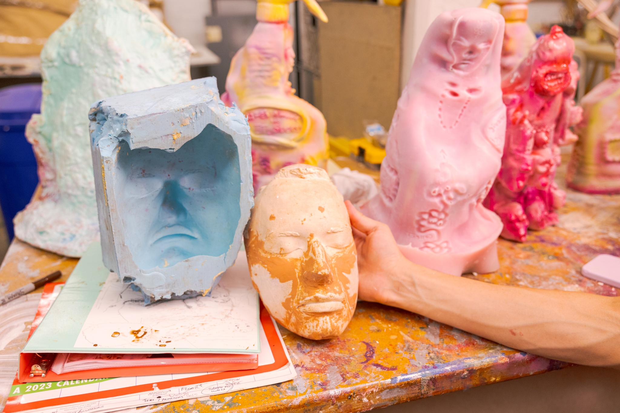 A collection of 3-D printed plastic objects sit on a wooden work table. Jake Brush reaches his hand over the table to hold up a cast of a face in orange plastic. 