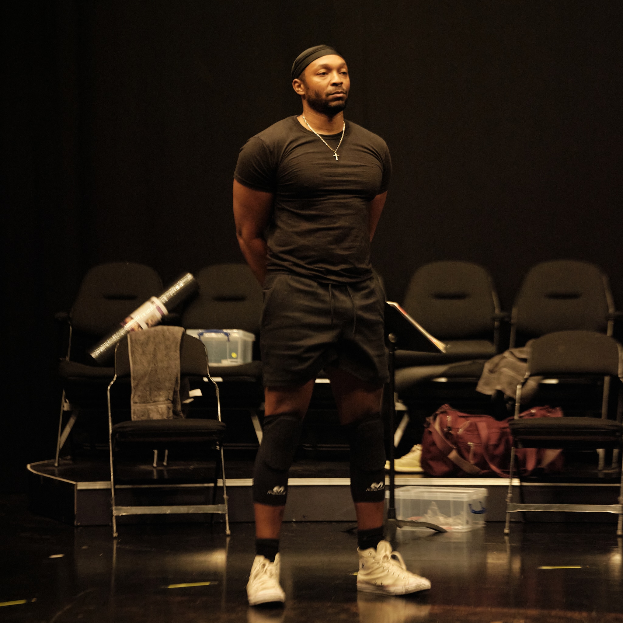 Actor Malcolm Mays, a Black man, stands in a black-box rehearsal studio. His legs are firmly planted and he stands straight with shoulders back, arms behind his back. He looks straight ahead, out of frame, and wears black t-shirt, shorts, and do rag.