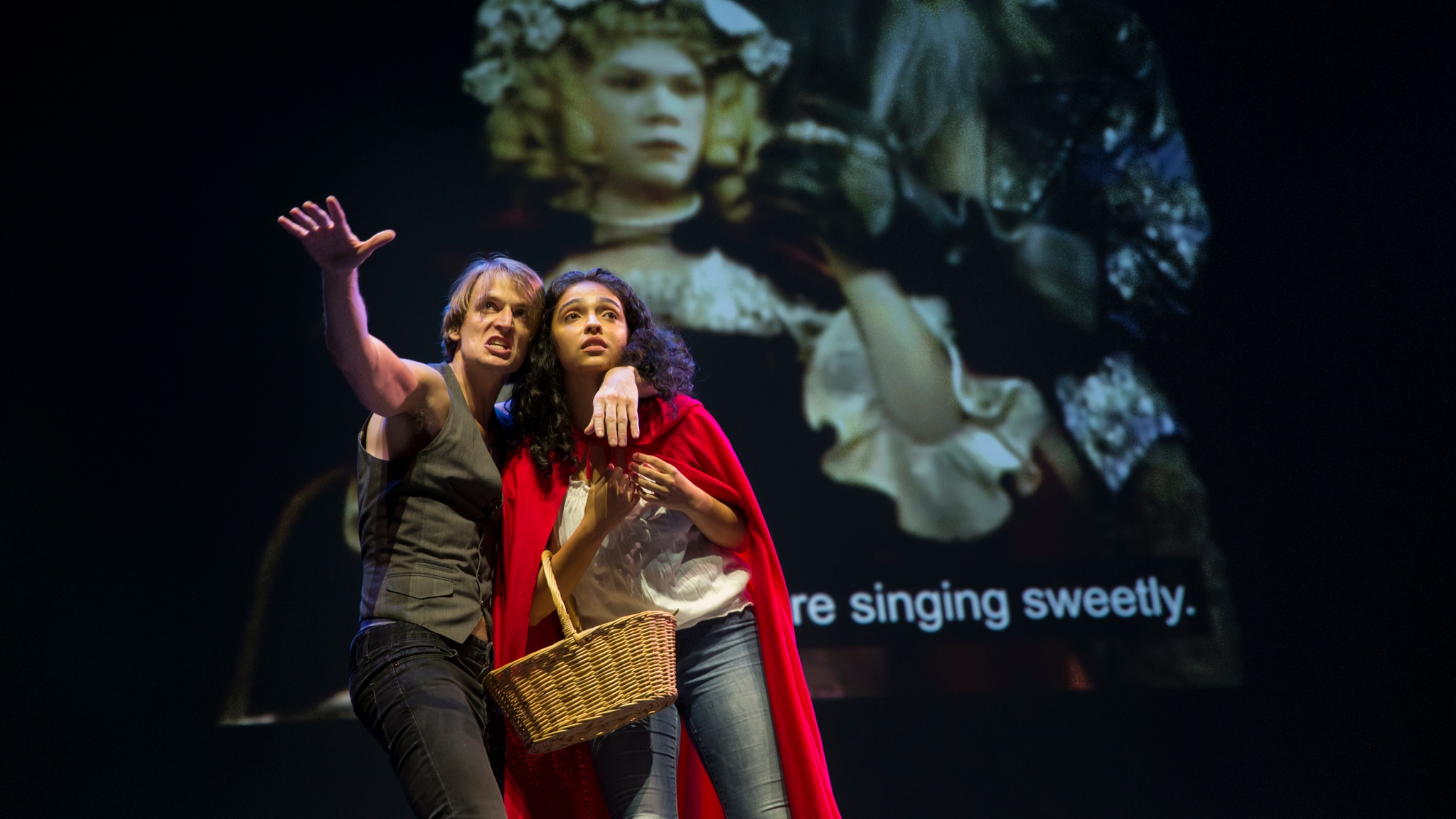 Two Deaf performers, a young man and woman, stand closely together on a stage with a video projection behind them. The white man, wearing a dark gray vest, has one arm around the shoulders and neck of the other performer while stretching his other arm out toward us. The young Hispanic woman, with curly dark hair to her shoulders, wears a red riding hood cloak and holds a basket in the crook of her arm. The video projection shows a period scene from a performance of Into the Woods, with the subtitled words “singing sweetly” visible behind the performers. 