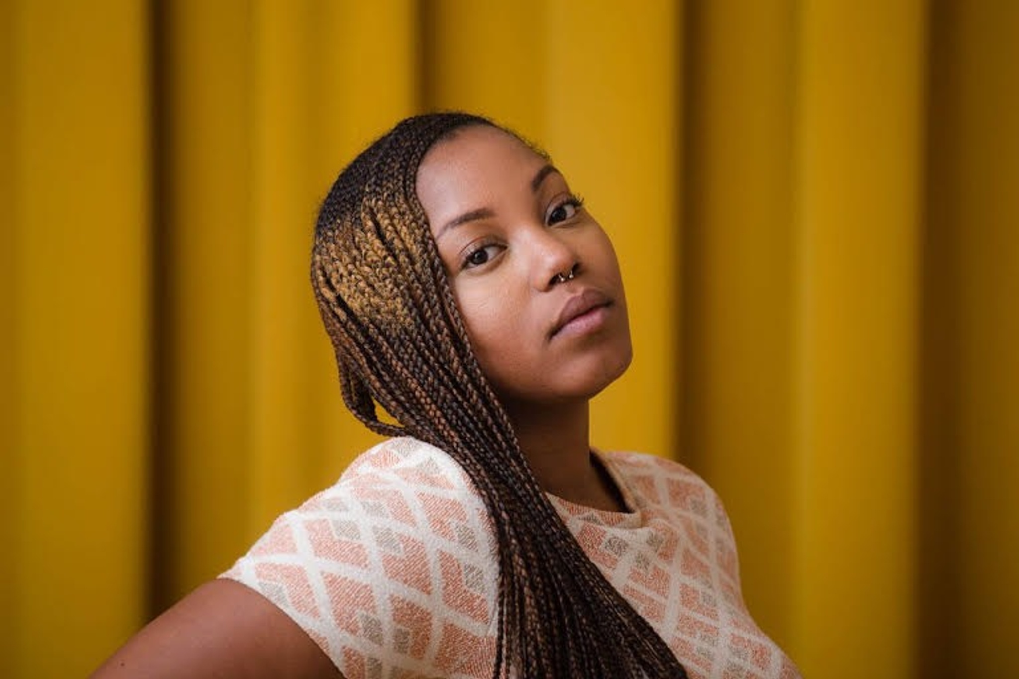 Poet Camonghne Felix poses in front of a gold curtain. Turned slightly to the side, she looks at the camera, has long braided hair that falls over her right shoulder, and wears a pink-and-gray, diamond-patterned shirt. 