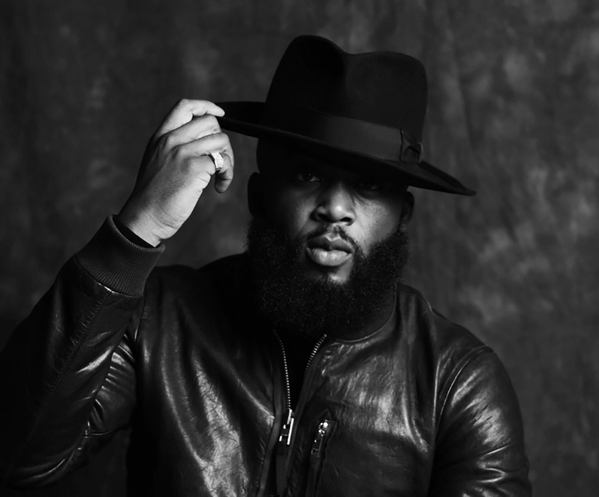 A black-and-white photo of Julien ‘OG’ Richardson against a studio fabric background. OG wears a leather jacket and holds one hand up to the wide brim of a hat he is wearing. Light falls on the left side of his face, leaving the right side in shadow. 