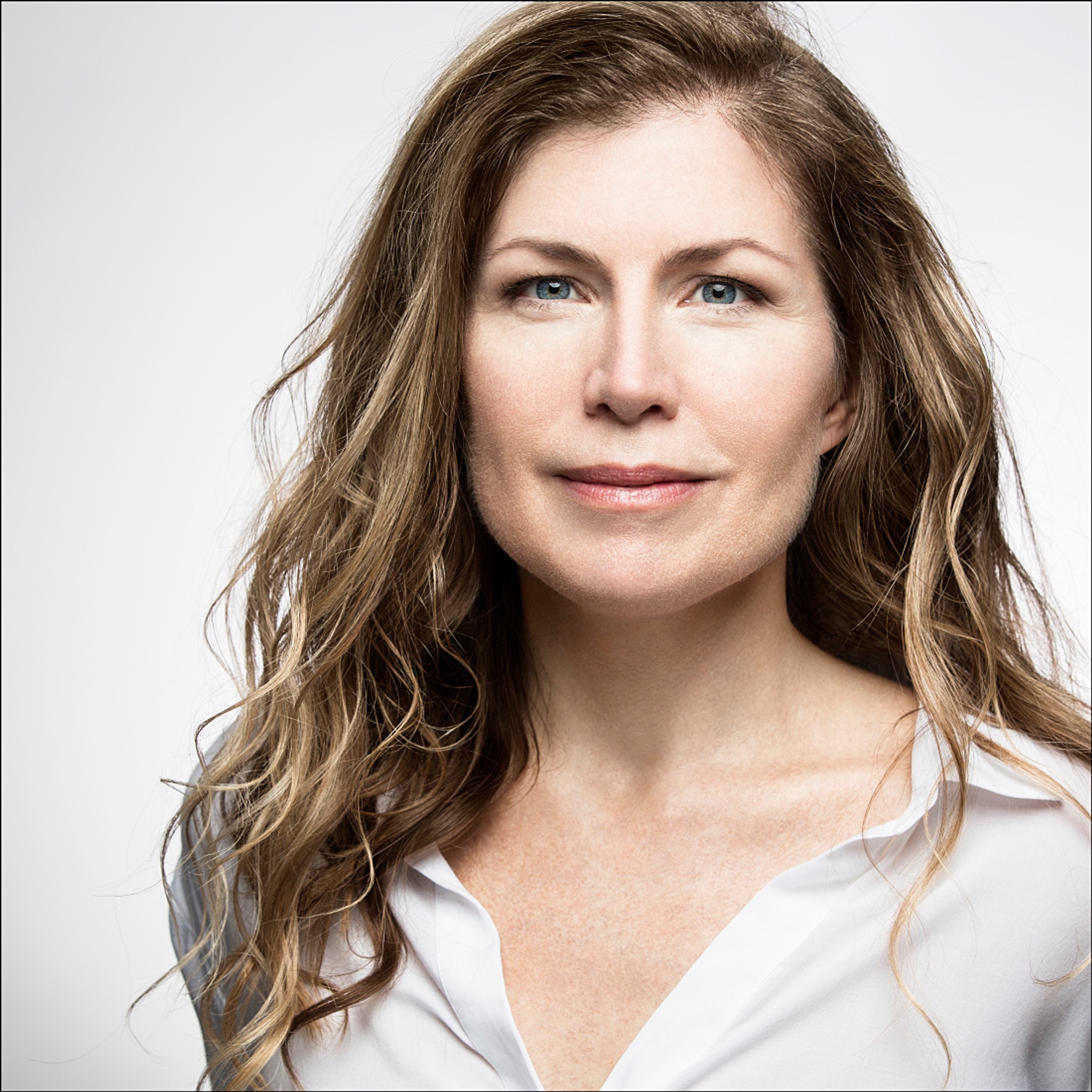 A portrait of actor Mary Stillwaggon Stewart, a white woman with long wavy light brown hair. She looks intently at the camera against a white background.