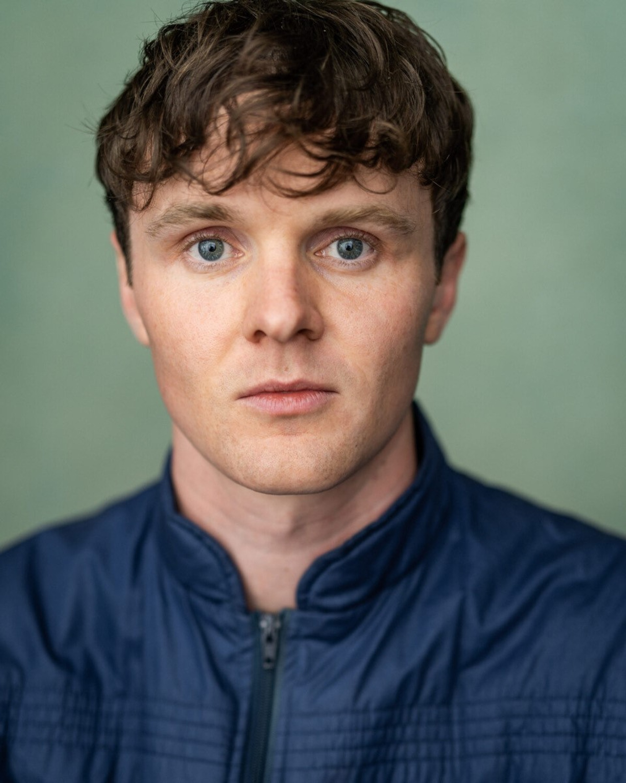 A headshot of actor Doug Colling, who looks directly at us. He is white and has light brown hair that is styled to fall down over his forehead. He has blue eyes and wears a blue jacket with a stand collar. 