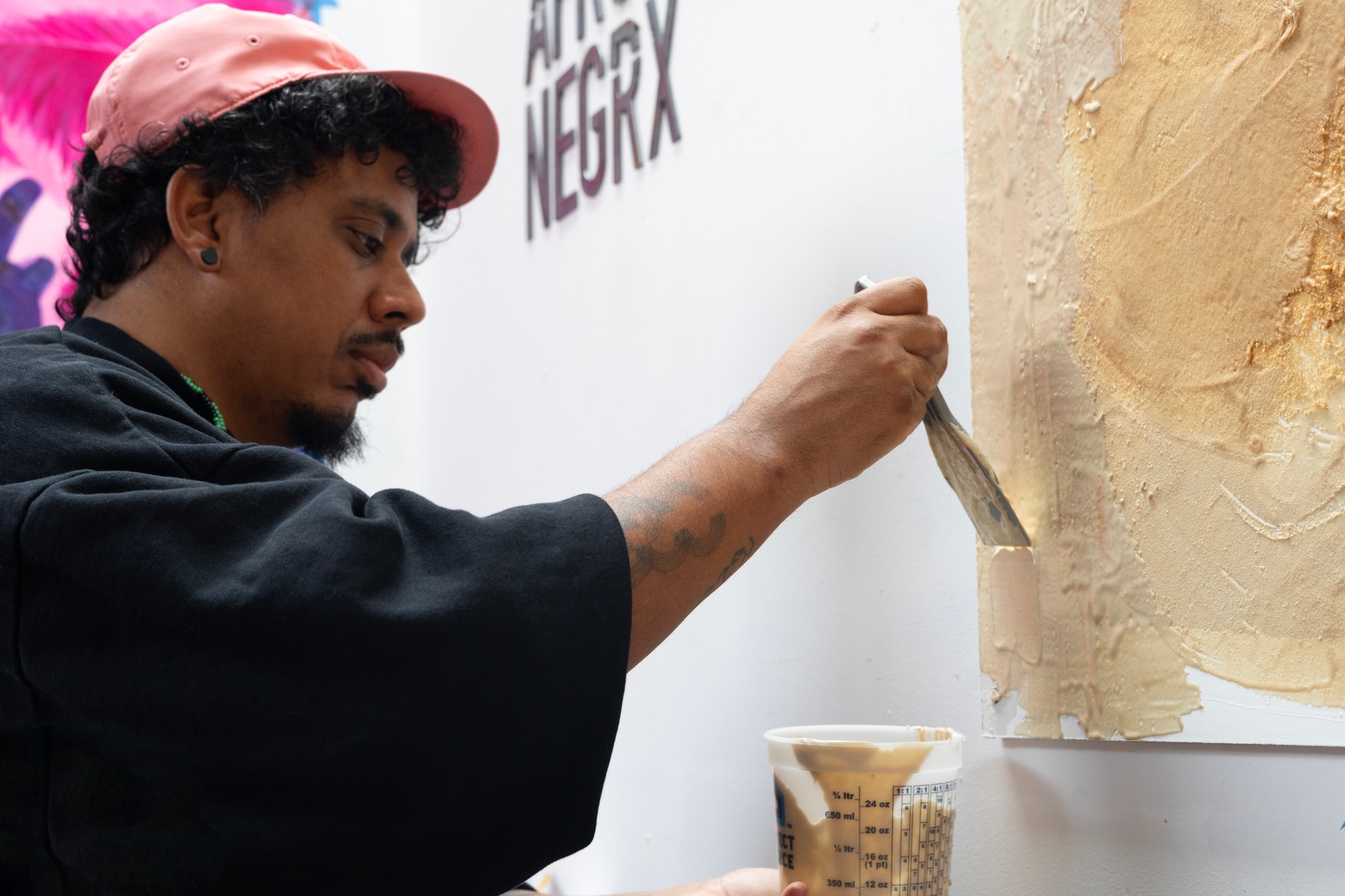 Artist Luis Vasquez La Roche works on a painting in his studio. He swipes a paintbrush upward on a white canvas hanging on the wall in front of him. The painting is in warm tones of yellow and brown and seems to be more of an underlayer than a representational image. He looks intently at his work. 