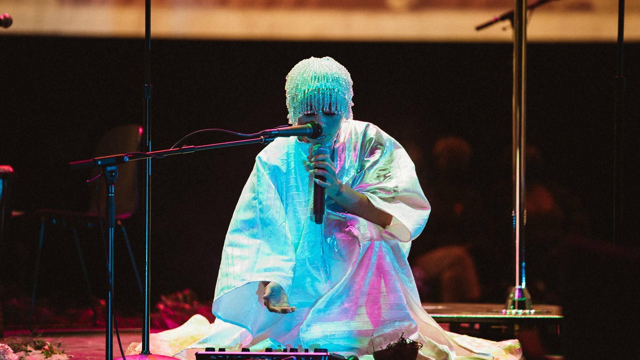 A performer sits on a stage with a microphone held to her face. She wears a white dress that engulfs her body and a shimmering, beaded cap on her head.