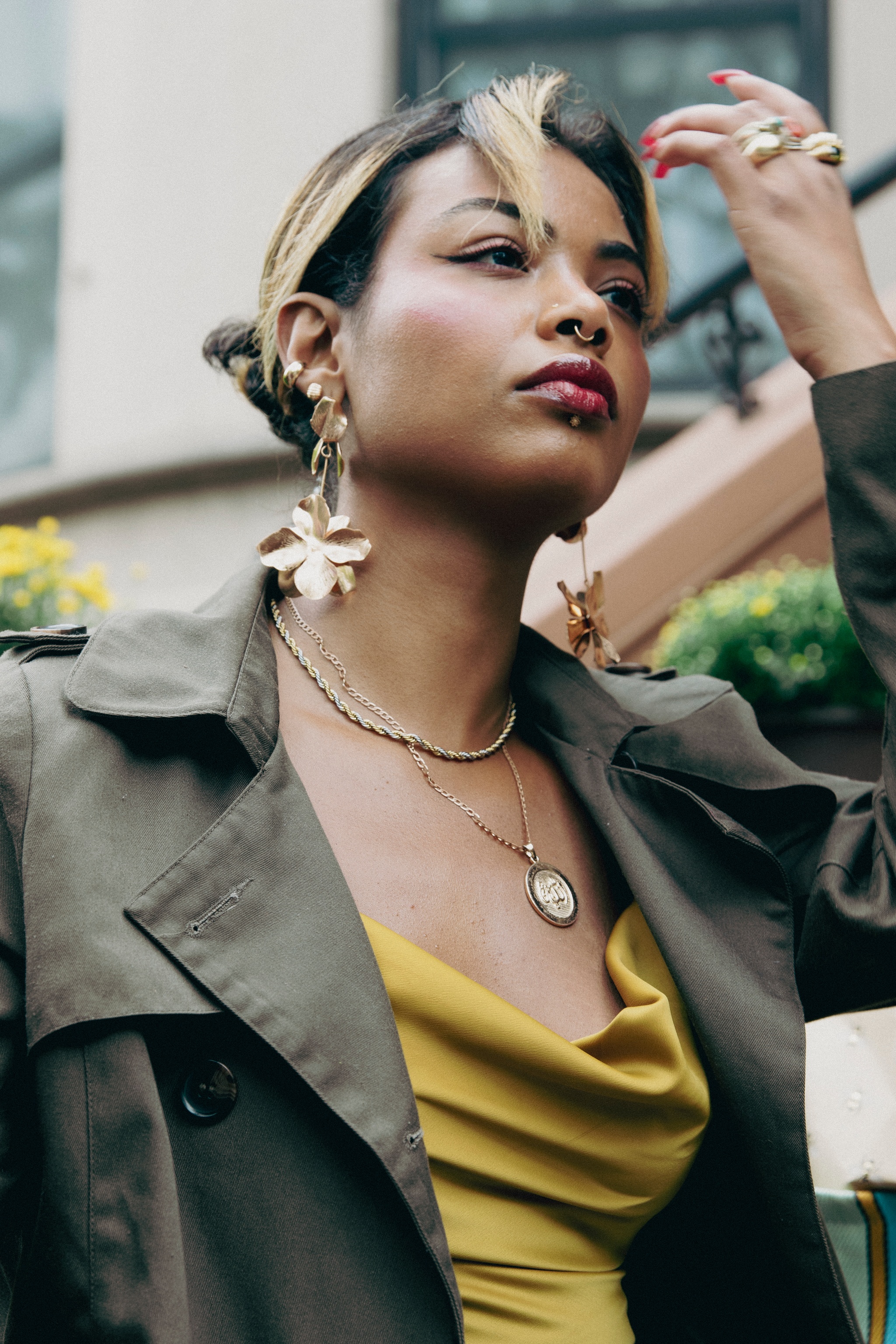 A portrait of artist Marwa Eltahir. Marwa is a Sudanese American woman who is seen outside in front of a brownstone building from an angle slightly below. She swipes windswept bangs from in front of her face.