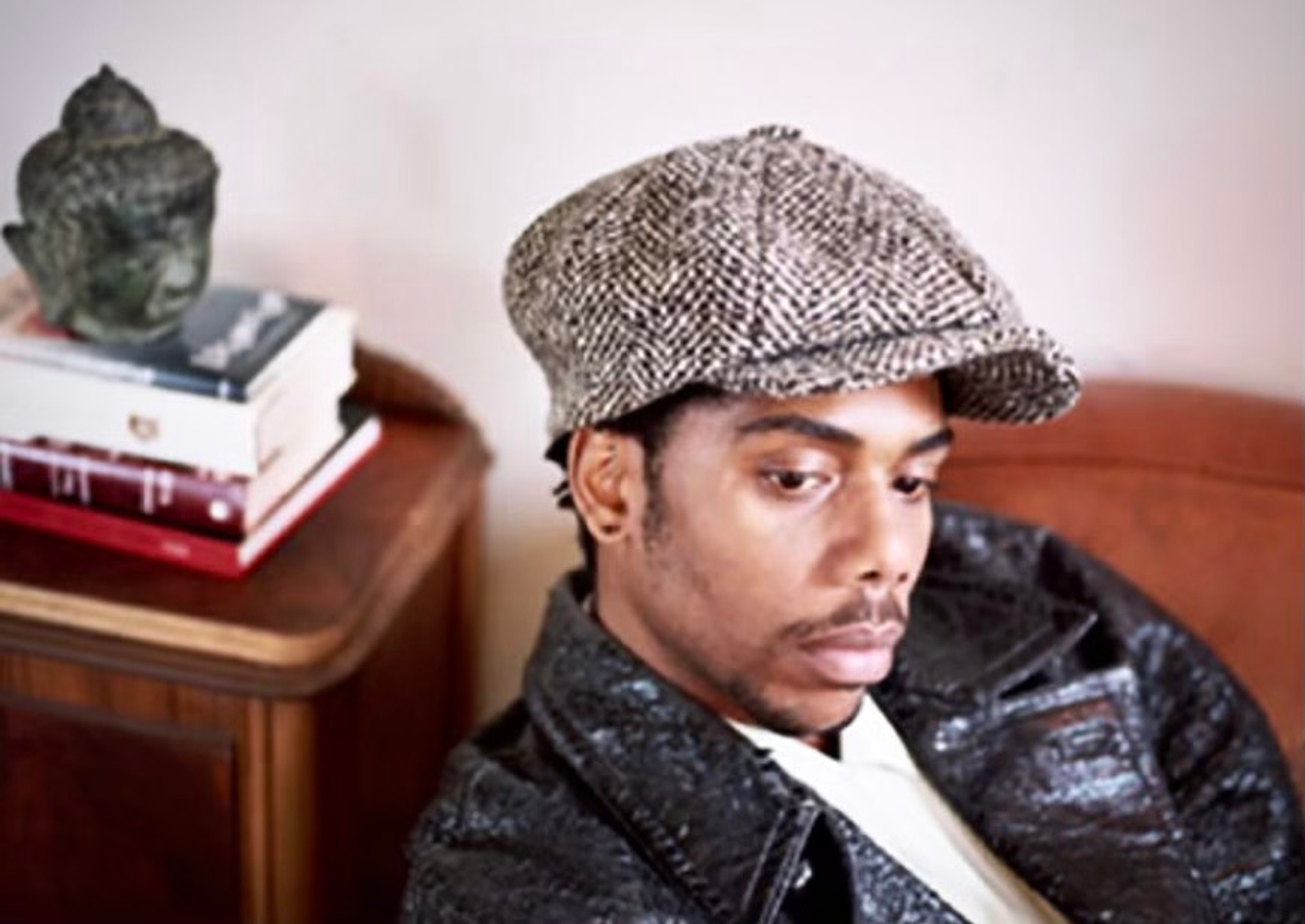 A portrait of Carl Hancock Rux, who is seen from an angle slightly above his head. He wears a leather jacket and an ivy cap. Behind his head is a stack of books with a Buddha's head sculpture atop it. 