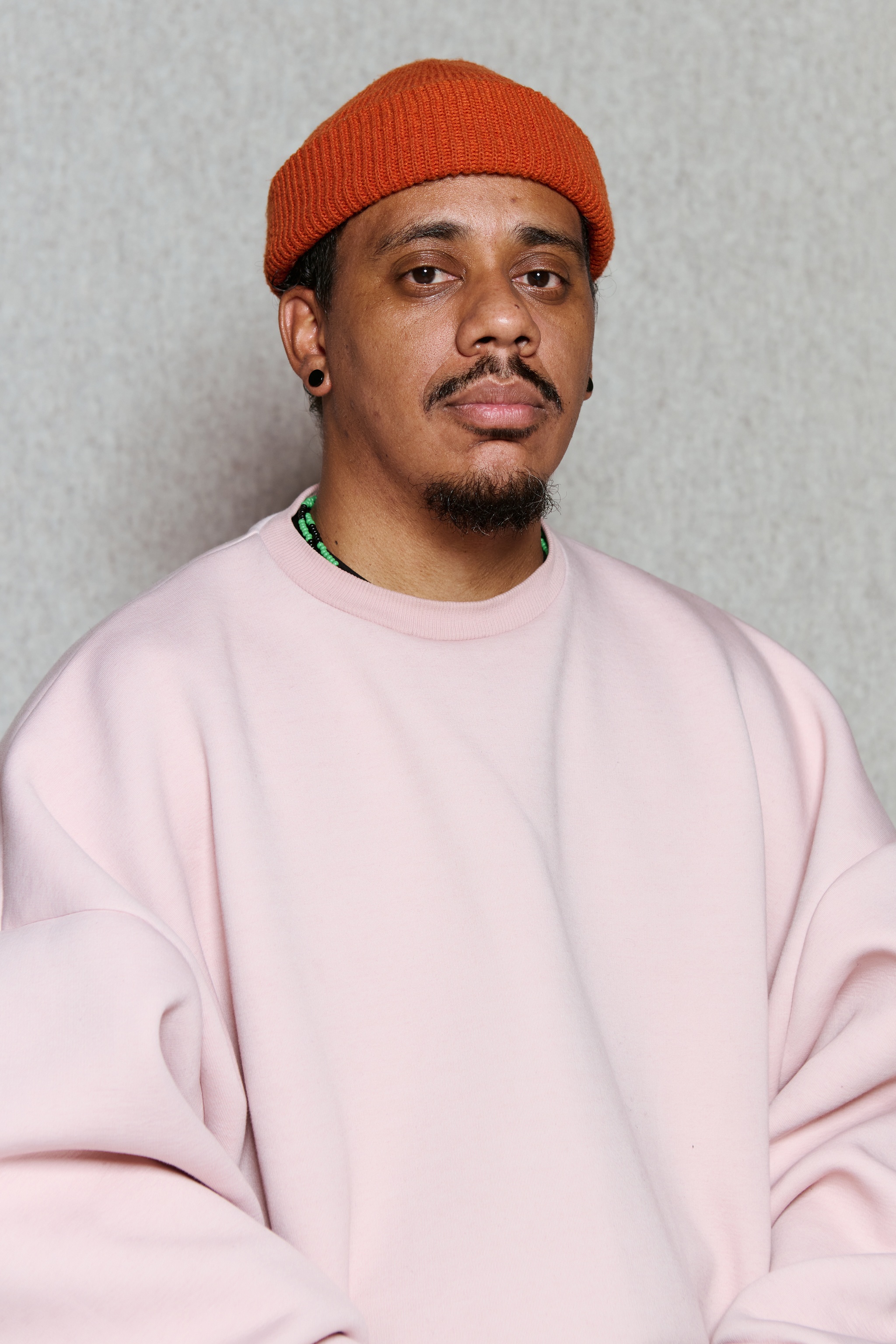 A portrait of artist Luis Vasquez La Roche. He has a mustache and goatee under his chin and wears a pink sweatshirt and burnt orange beanie. He poses against a beige felt background and looks directly at us with a calm, concentrated look on his face. 