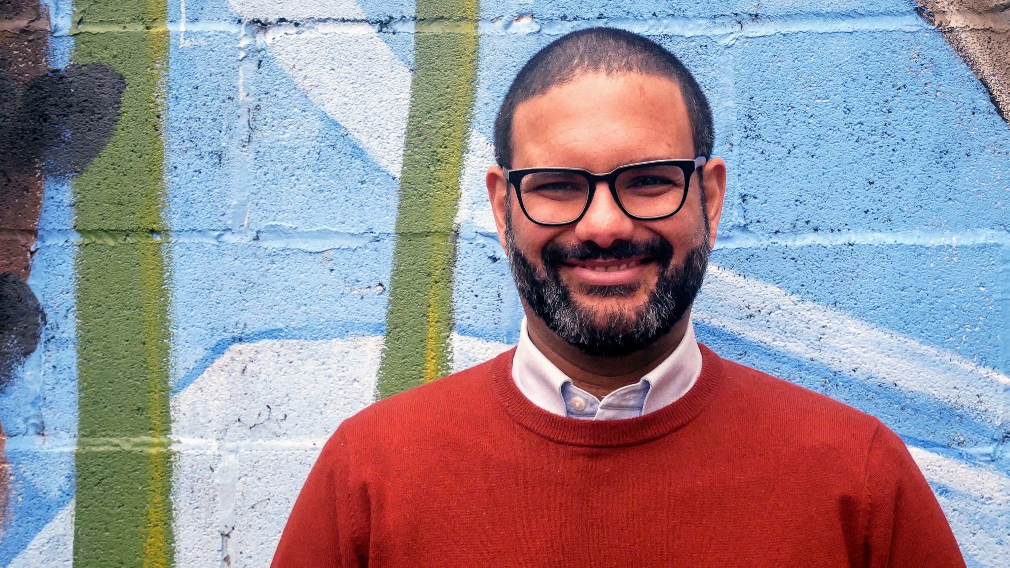 A photo of Hatuey Ramos-Fermín wearing a red sweater and glasses, standing against a mural painted on a cinderblock wall. 