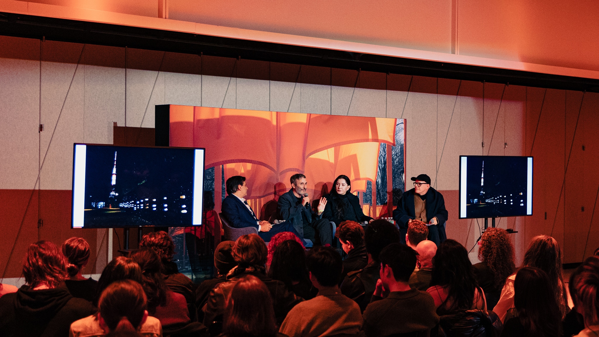 Four conversation panelists sit on a stage with a background behind them showing the saffron colored fabric of Christo and Jeanne-Claude's The Gates installation.