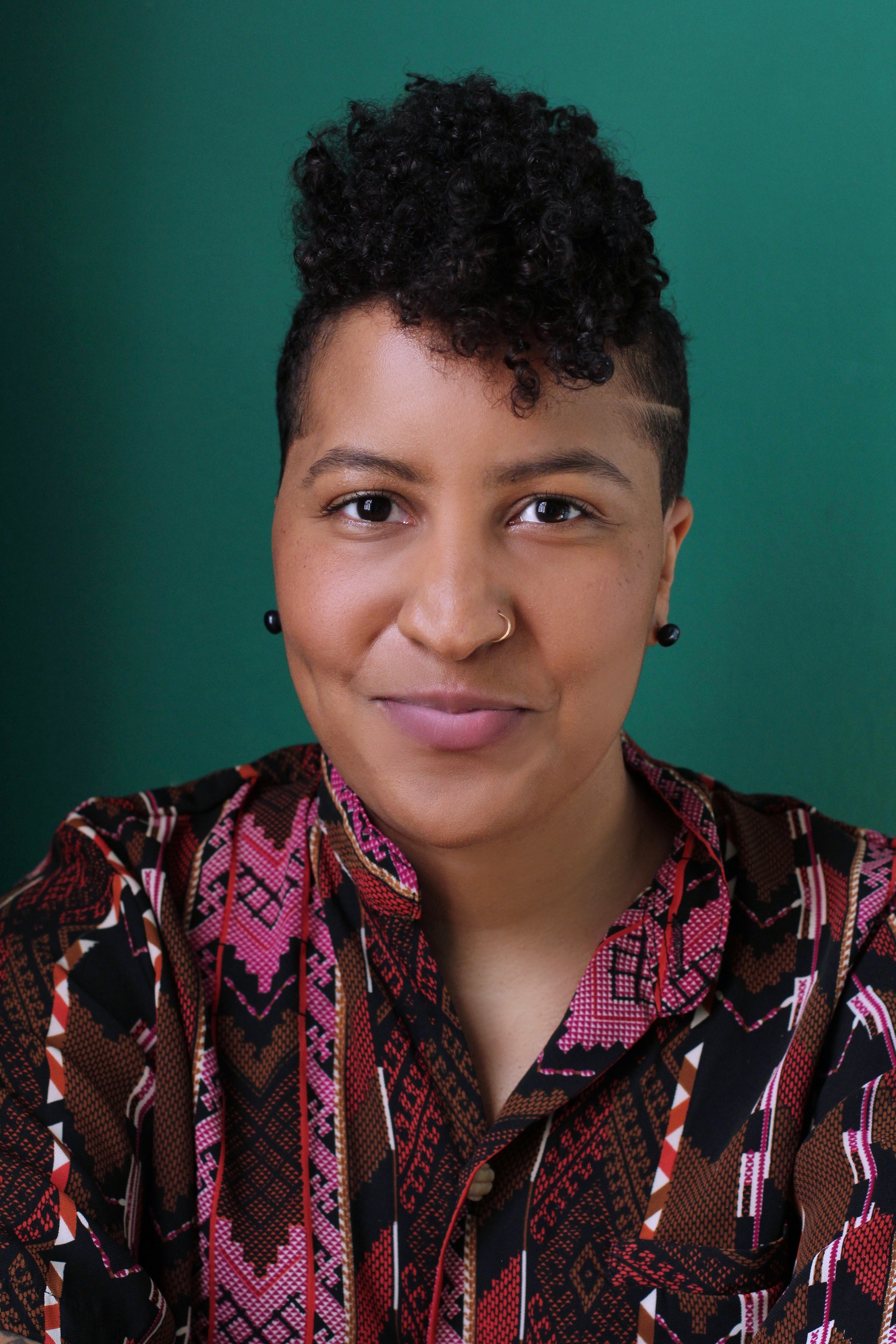 A portrait of Jordana De La Cruz who poses against a green background. She has curly hair that she wears high above her head with flat sides. She wears a print blouse in pink and brown tones and smirks playfully at us. Photo by Sammy Tunis