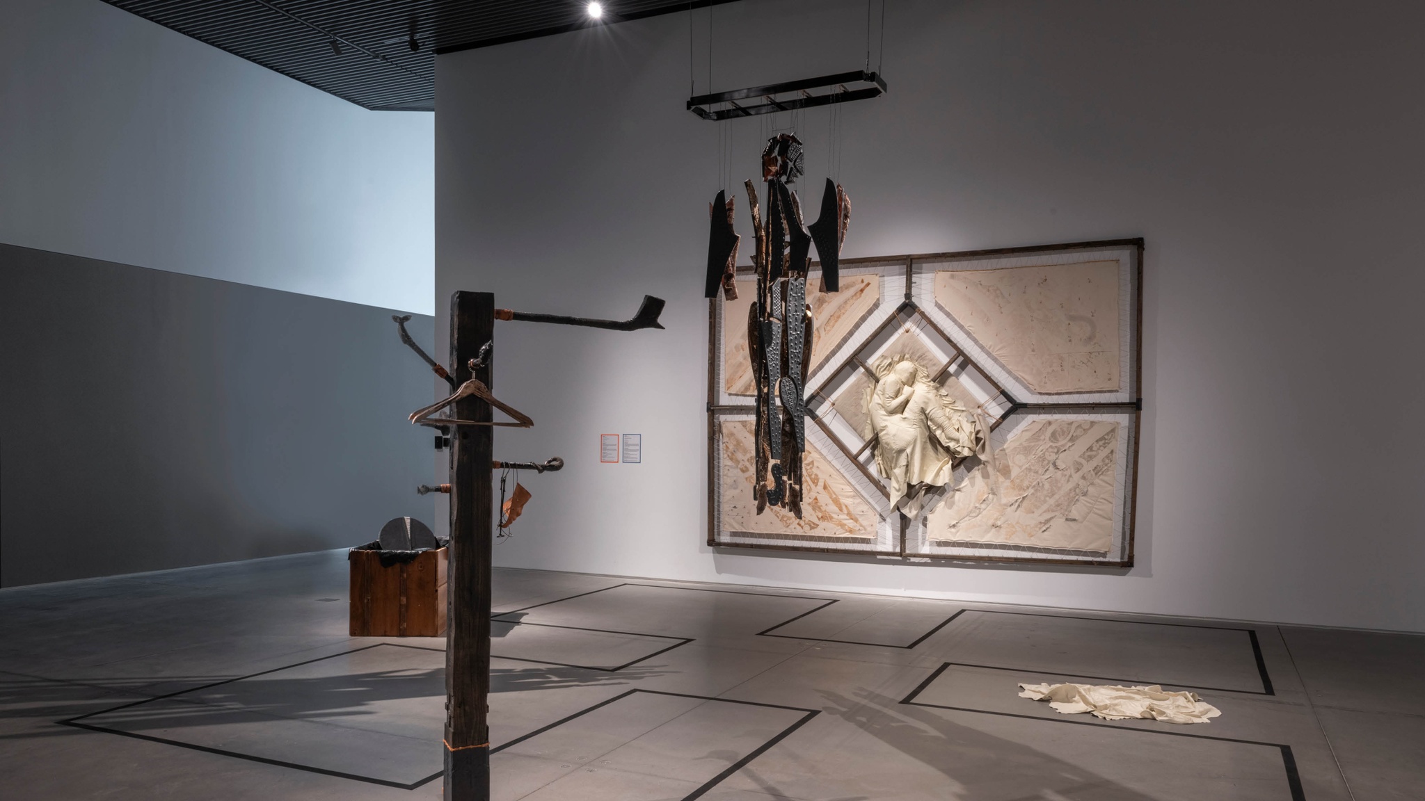 An installation of five artworks in a gallery. They are placed on the ground, hanging from a wall in the background, or hanging from the ceiling. The works on the ground include a box with leather scraps piled inside, a white piece of leather dropped on the floor, and a clothing rack similar to a coat rack. On the wall, a tapestry is stretched on a metal frame. At its center a human figure is formed out of white leather that has been stuffed. The figure is in the fetal position. Hanging at the center of these works is a pattern of the artist's body that has been cut out of black leather and affixed to shaped wood panels. The individual parts are attached by wires similar to a skeleton. 