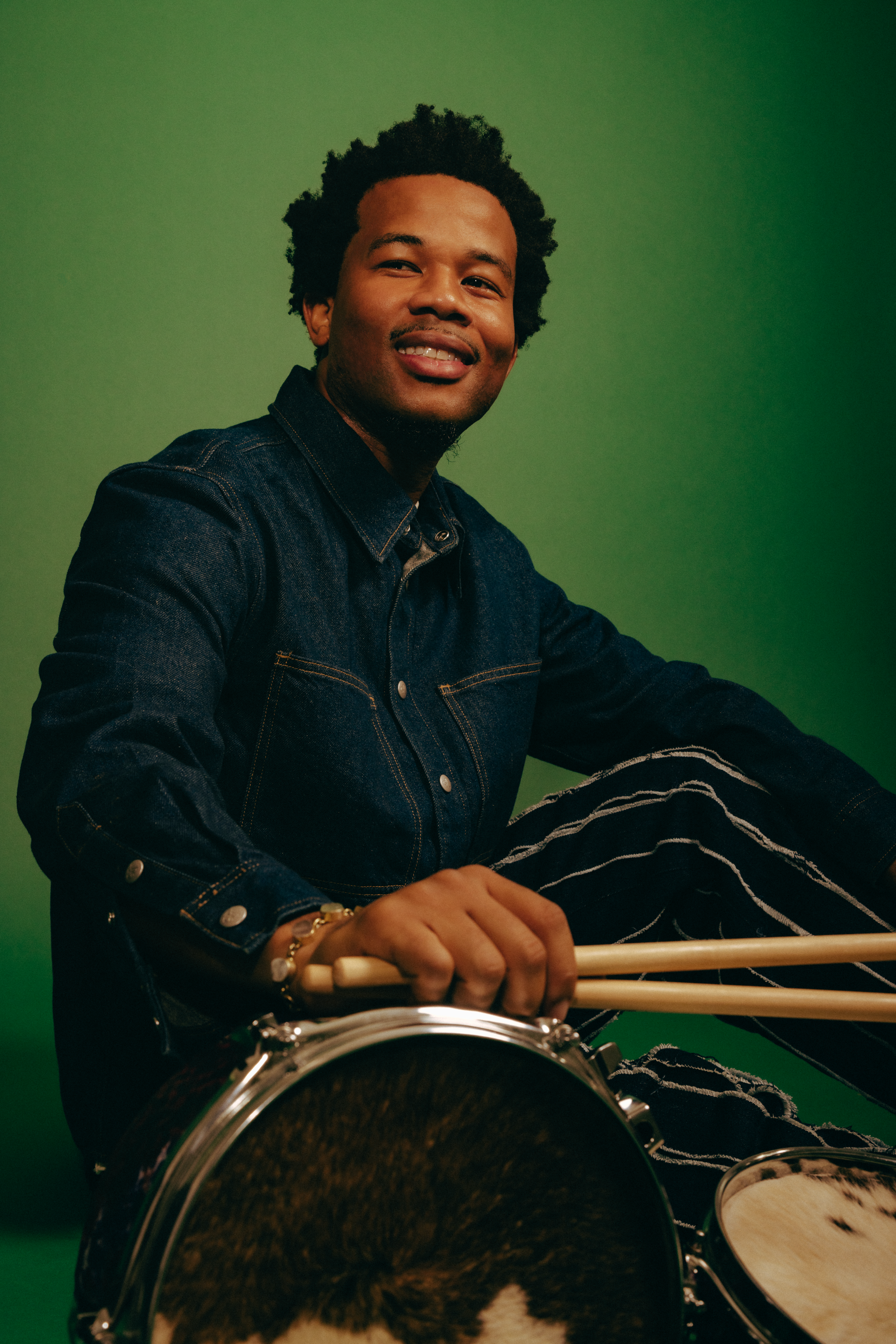 A studio portrait of Munir Zakee in front of a green backdrop. Munir, a Black man, smiles past the camera. He's wearing a denim jacket and striped pants. He's holding drumsticks in his hand which is resting on the edge of a drum.
