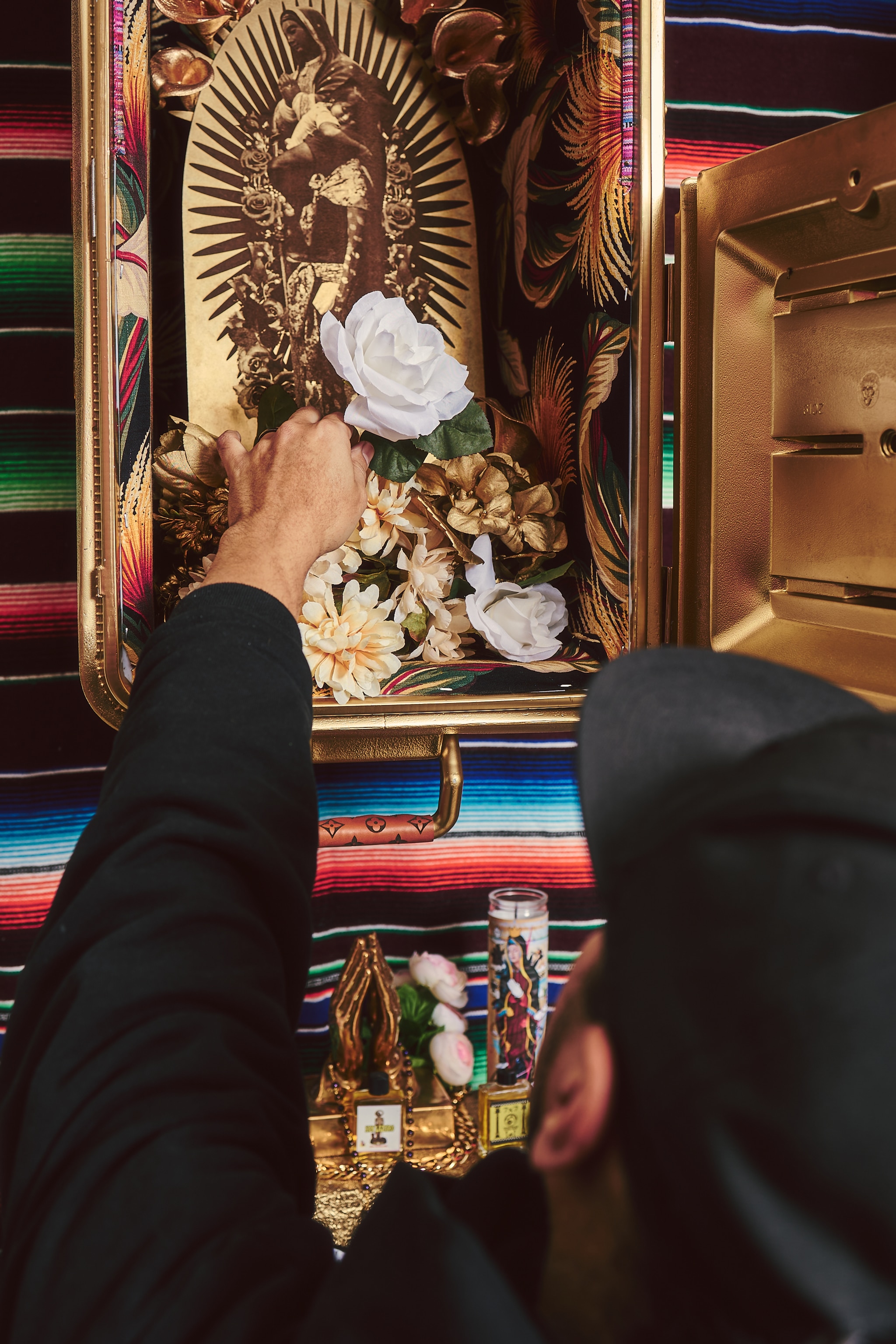 Artist Marka27 is seen from behind reaching up into an open ice cooler that has been painted gold. Inside is a devotional altar on which he reaches to pose a white flower. 