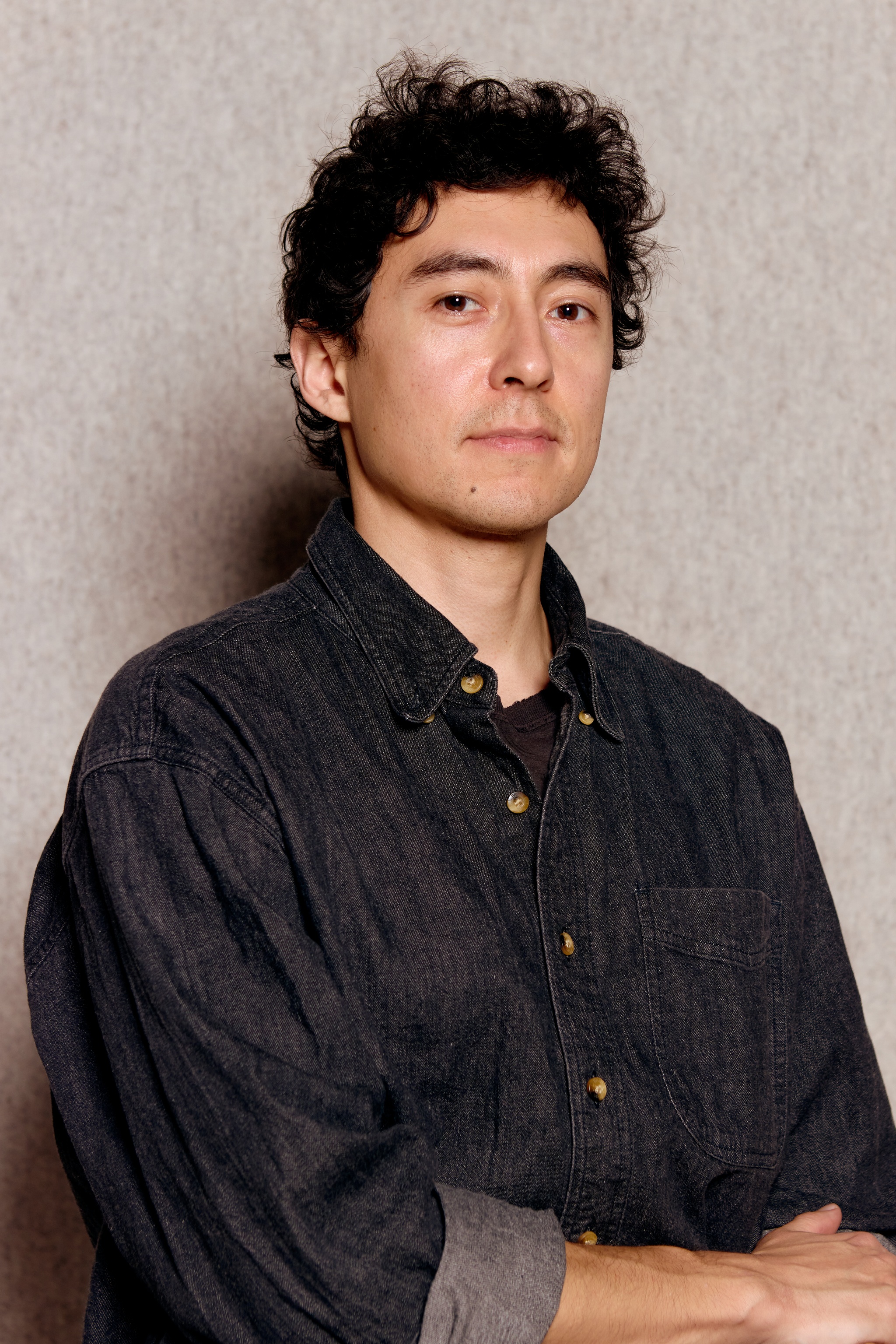 A portrait of artist Tyson Houseman. He sits in a denim button down shirt against a beige felt background. His arms are crossed at his waist at the bottom of the frame and he looks directly at us, with eyebrows slightly raised. 