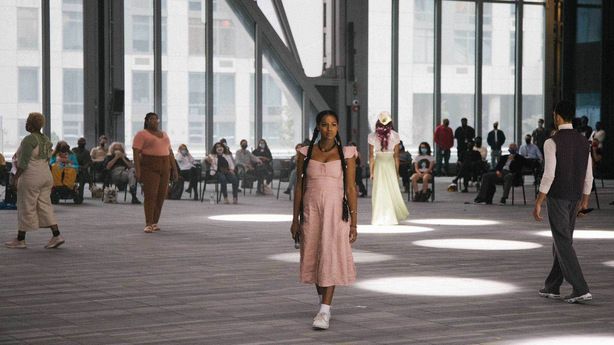 Five performers in a large performance space walk in different directions from one another. In the background, the audience is seated in pairs. 