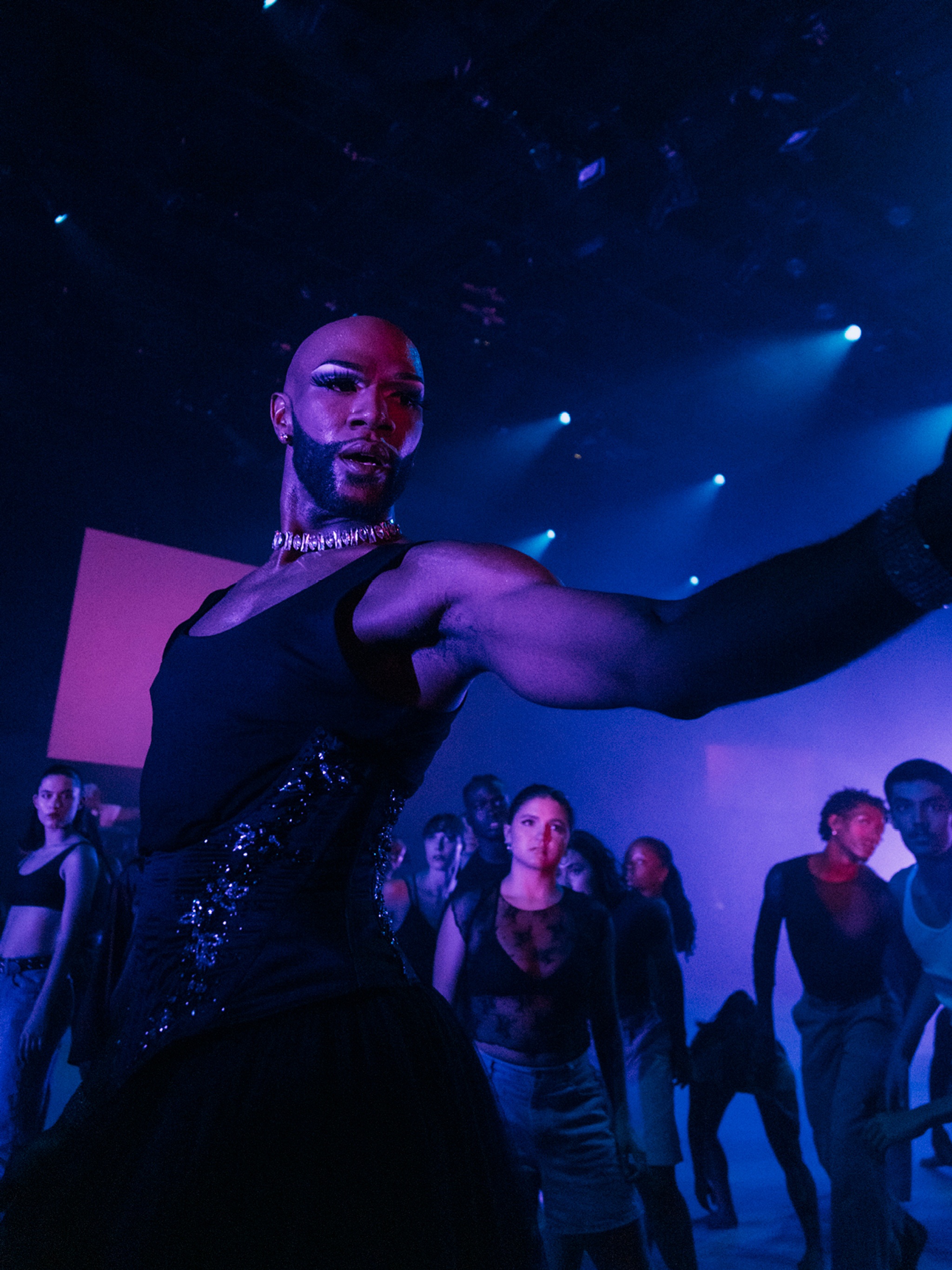 Cain Coleman, a Black performer and artist, dances with other dancers in "New Information." Cain wears a black gown and choker, and stretches one arm out as he looks to the side. Behind him in the space lit with purple light, dance numerous other dancers. 