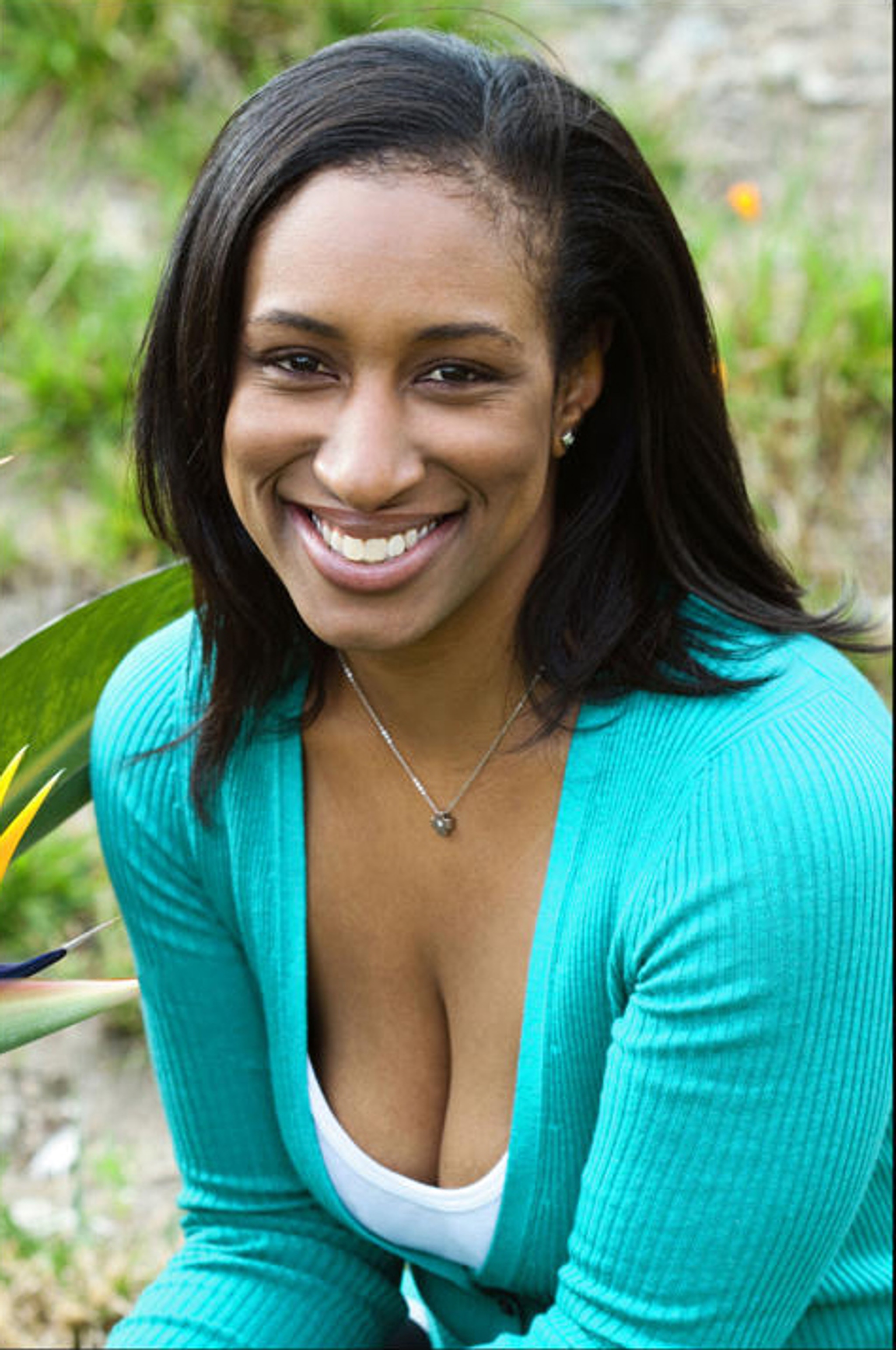 A portrait of performer Khalilah Smith who poses outside with green foliage blurry in the backgroud. Khalilah is a Black woman with hair parted to the side that falls to her shoulders. She smiles happily at us and wears a turquoise long sleeve shirt with a white tank top underneath. 