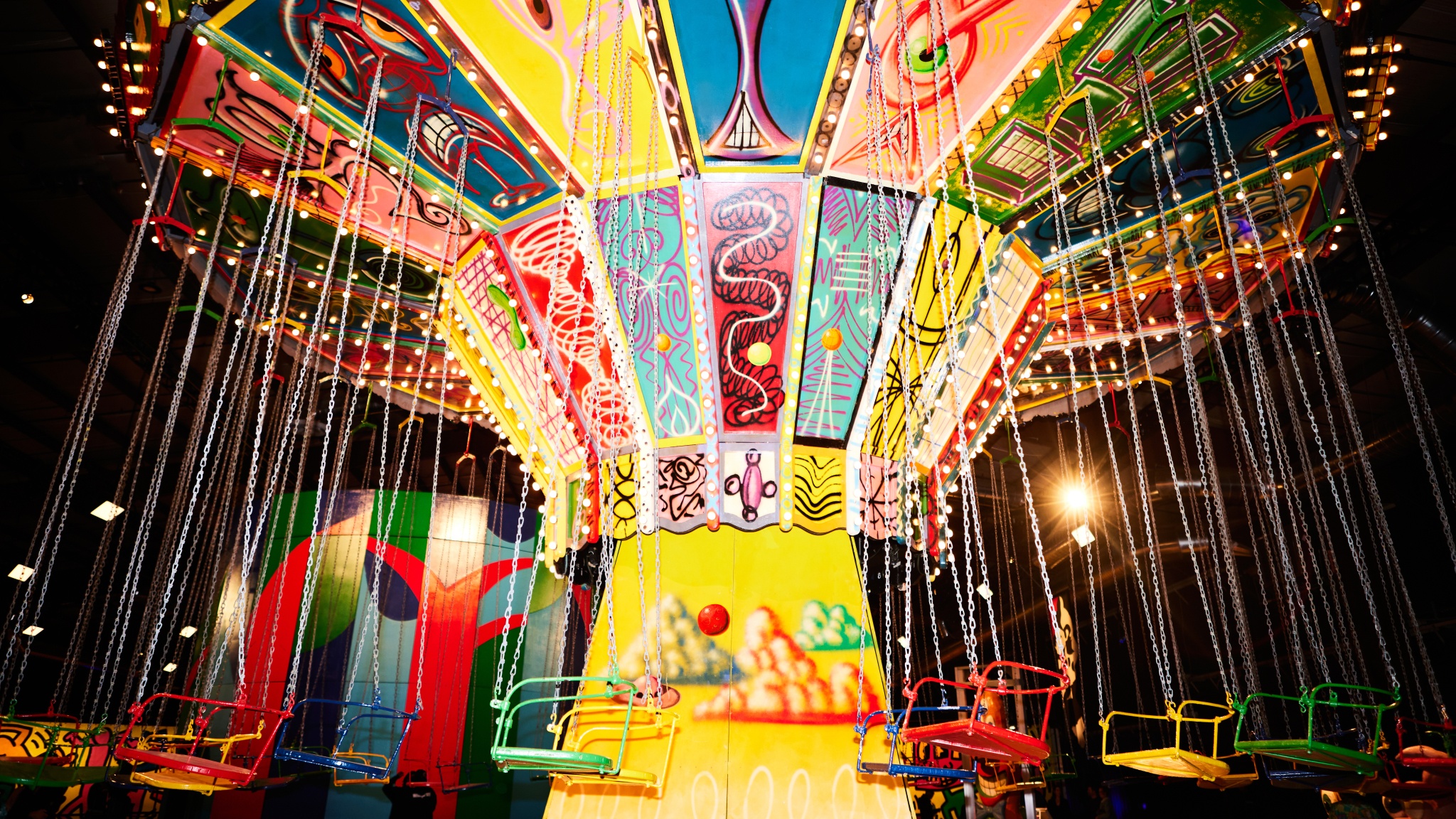 A carnival chair swing ride by artist Kenny Scharf with colorful panels designed by the artist is illuminated by bright light bulbs on the ride. Its chairs in different colors hang from chains around it. 