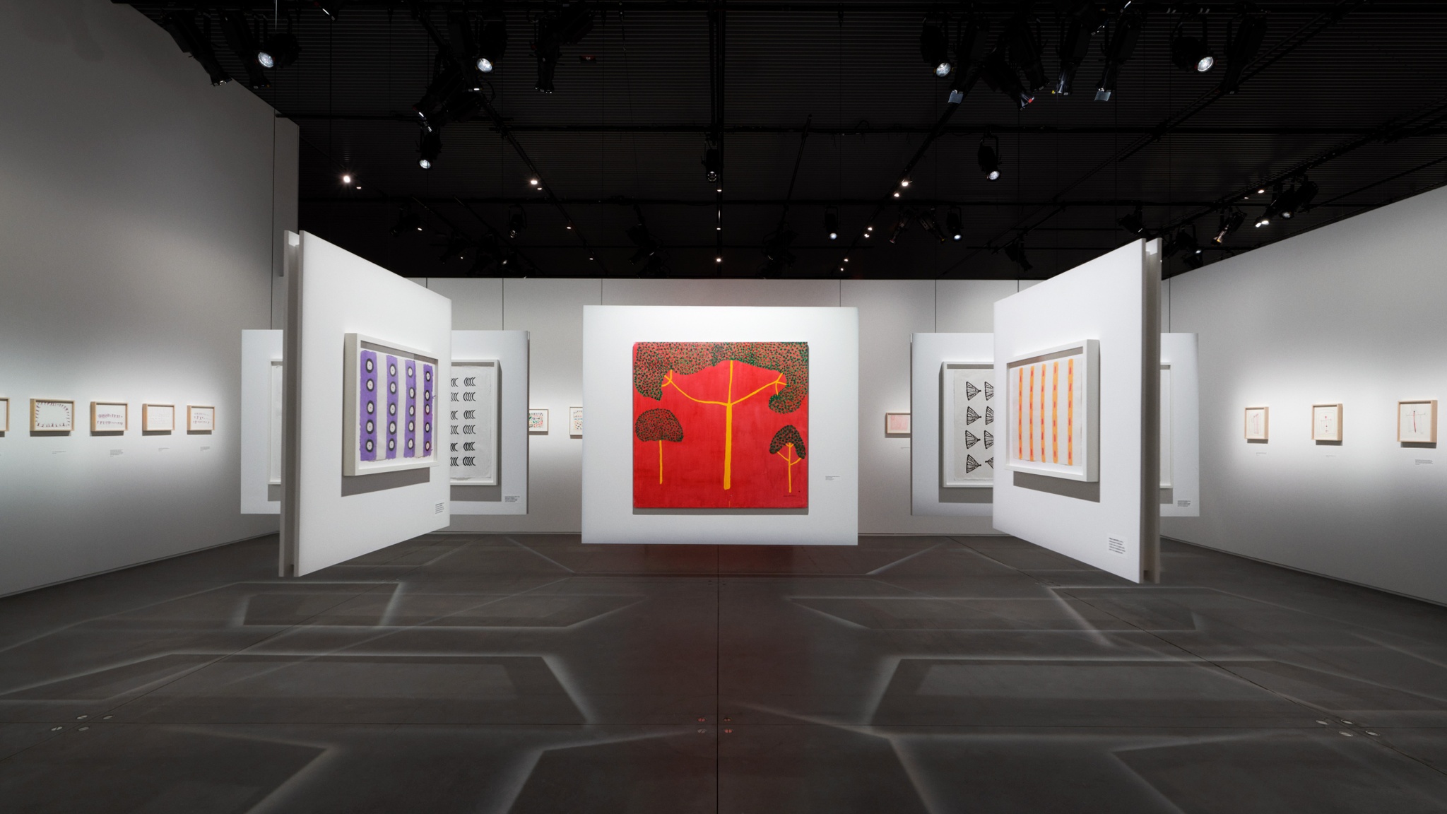 An art gallery with drawings and paintings on walls suspended from the ceiling and on fixed walls. The suspended walls make shadows of intersecting lines on the gray concrete floors. Situated prominently in the center of the gallery is a painting of three trees with yellow trunks and green canopies against a bright red background.