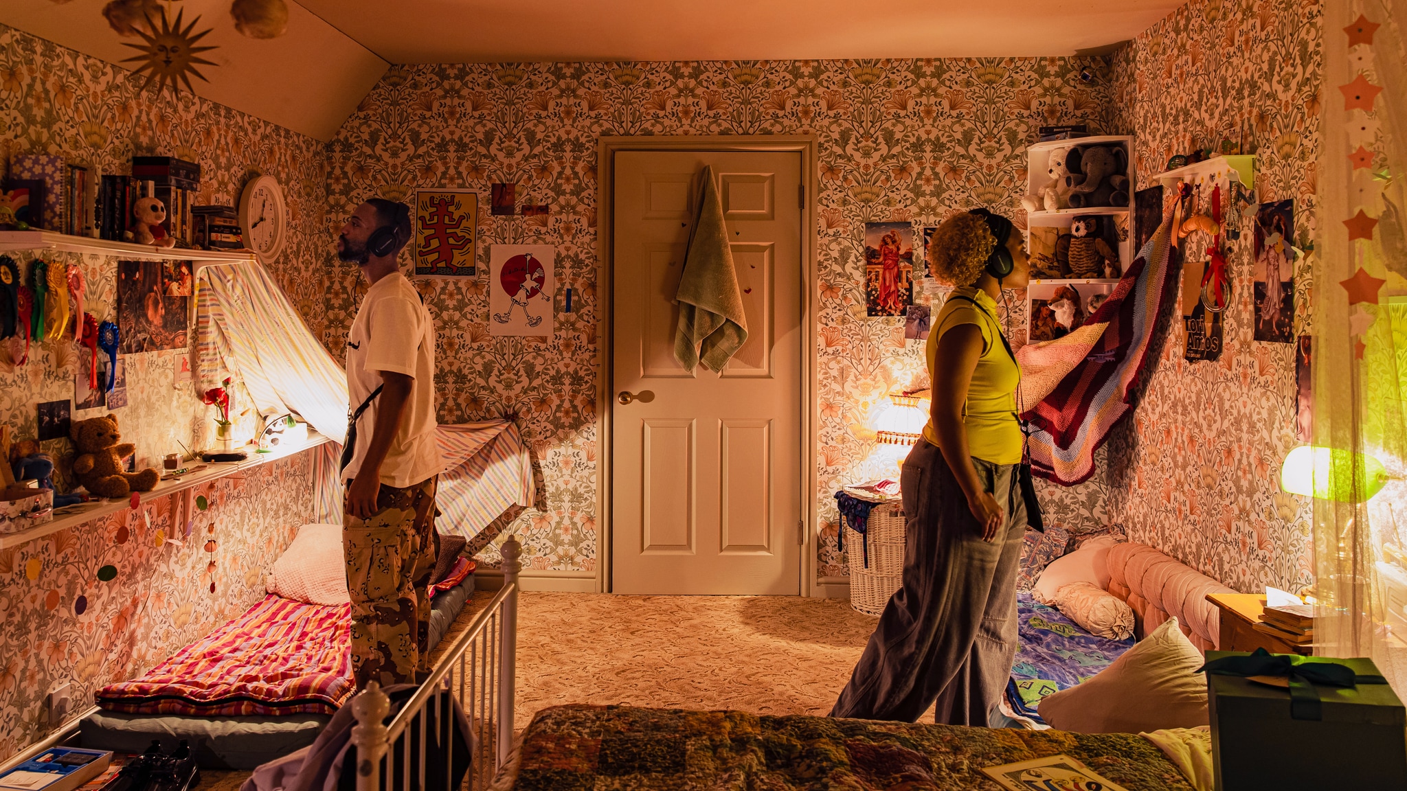 Two Viola's Room audience members stand in a teenage girl's bedroom from the 1990s. They wear headphones and stand facing away from each other, inspecting the objects in the wallpapered room, including movie posters, books, and other objects. 