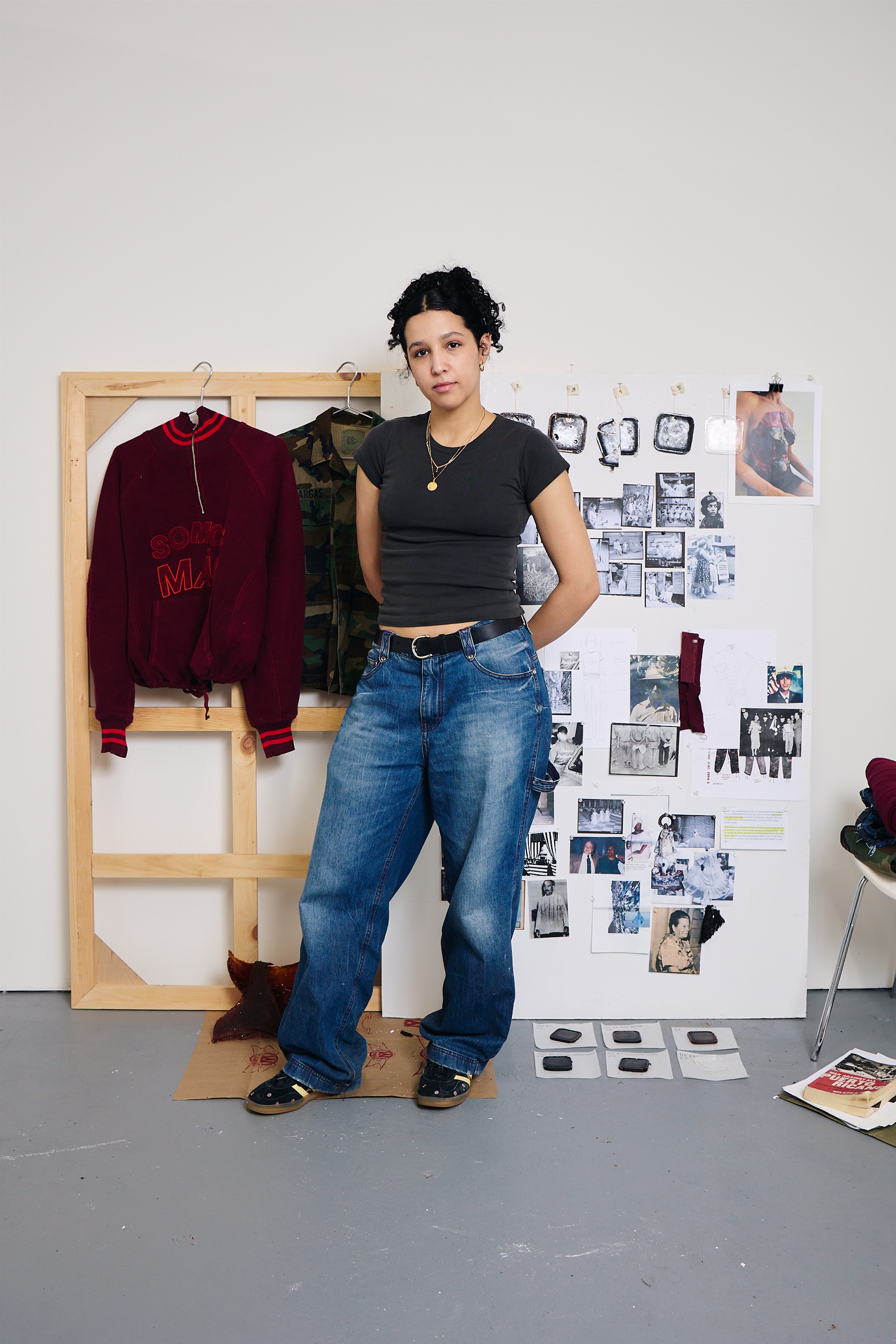 The artist Mel Corchado poses in her studio in front of clothing including a red jacket hanging from a wooden frame behind her. She stands with hands behind back, looking at us with a slight smile. She has curly dark hair pulled back behind her head, wears a pair of baggy jeans, and a fitted dark gray t-shirt. 