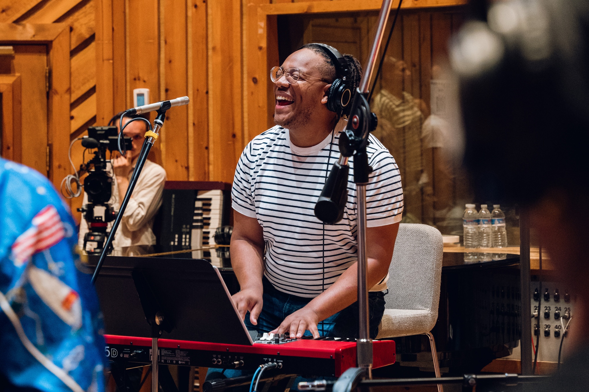 Troy Anthony, a Black composer and musician, stands in a wood-lined recording studio surrounded by mic stands and singers. He his laughing joyously as he plays a keyboard, wearing a striped t-shirt and headphones