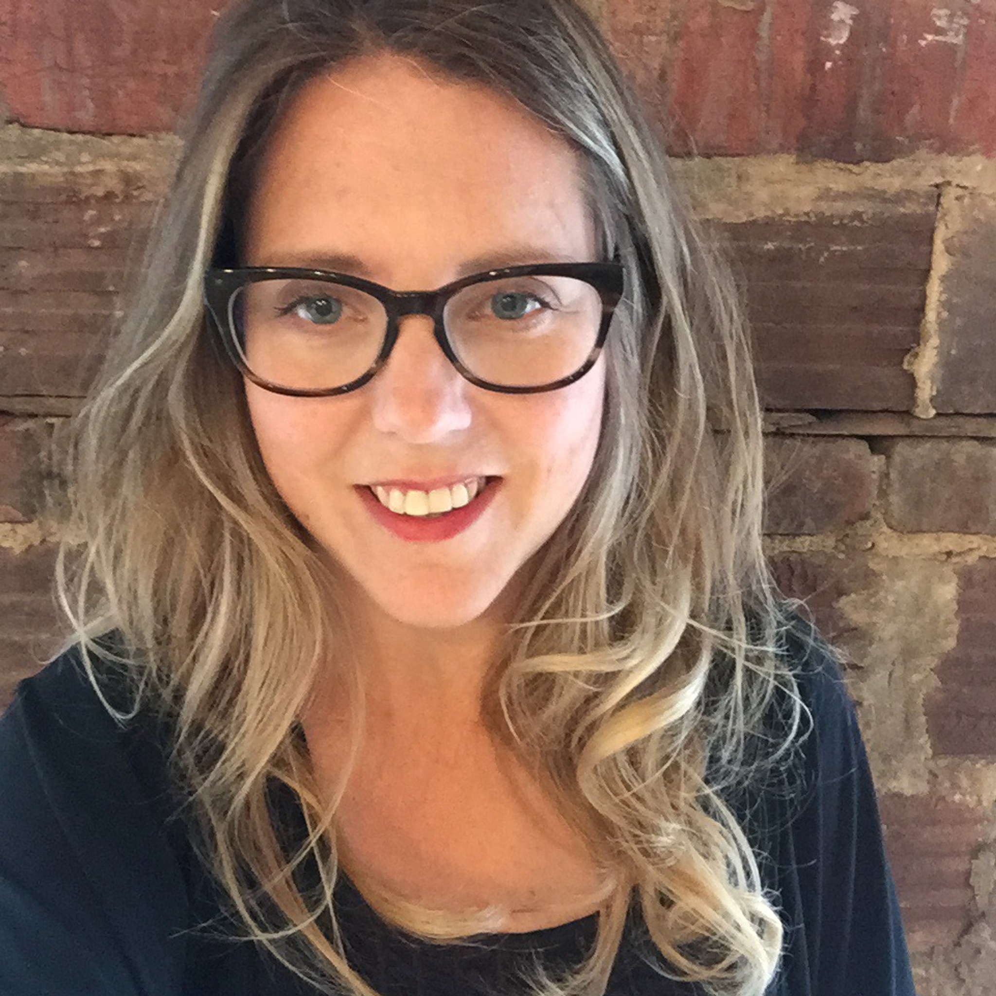 A photo of Andria Hickey, who wears glasses, smiles into the camera, and has hair falling over her shoulders. 