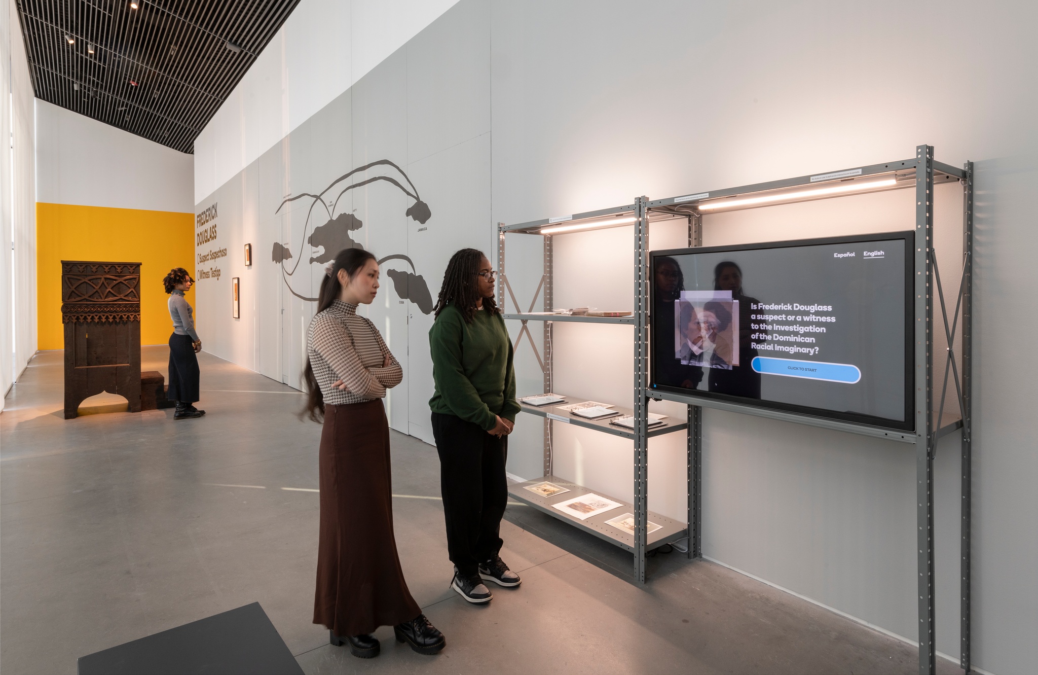 A long narrow gallery. Its long wall is painted gray and the intersecting wall at the end of the gallery is yellow. Two visitors stand in the foreground watching a tv screen on a metal shelf. In the background, another visitor looks at a print hanging on the wall. Beside her is an imposing pulpit, like you might find in a Gothic church. 