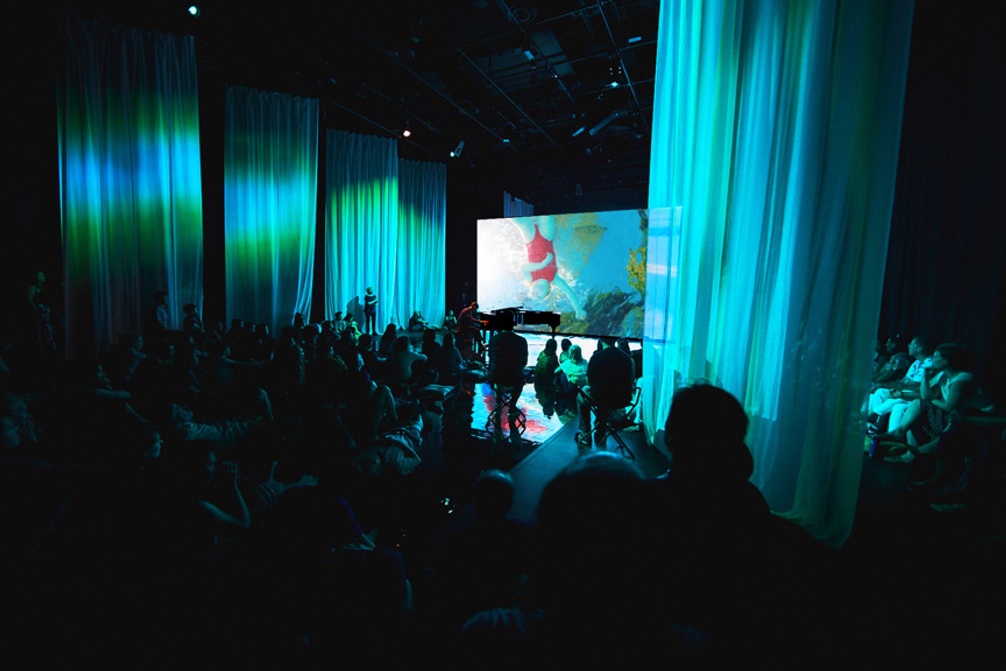 Audience viewing film on large screen in blue-lit space.Capture_315_002cc.jpg