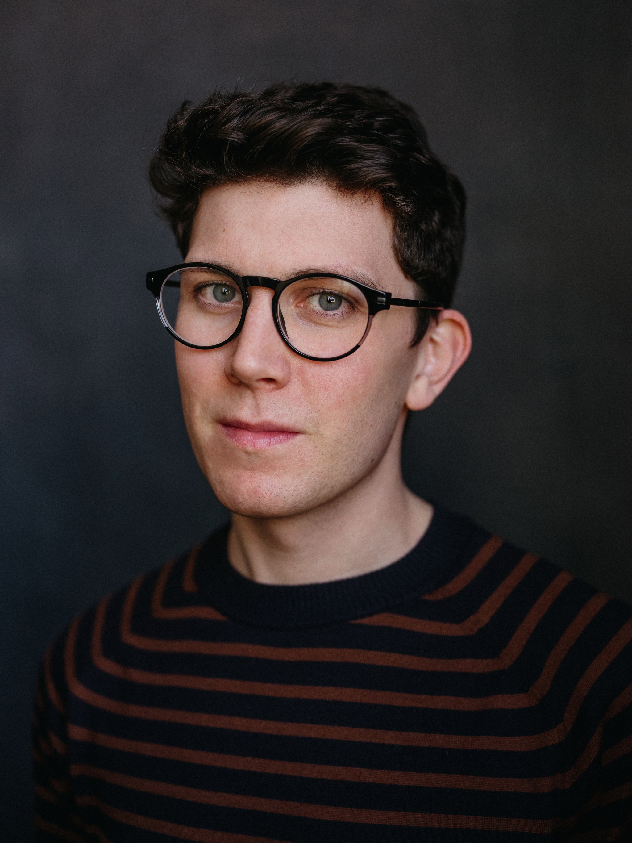 A portrait of actor Al Coppola, a white man with short brown hair wearing circular-framed glasses and a red and blue striped sweater. 