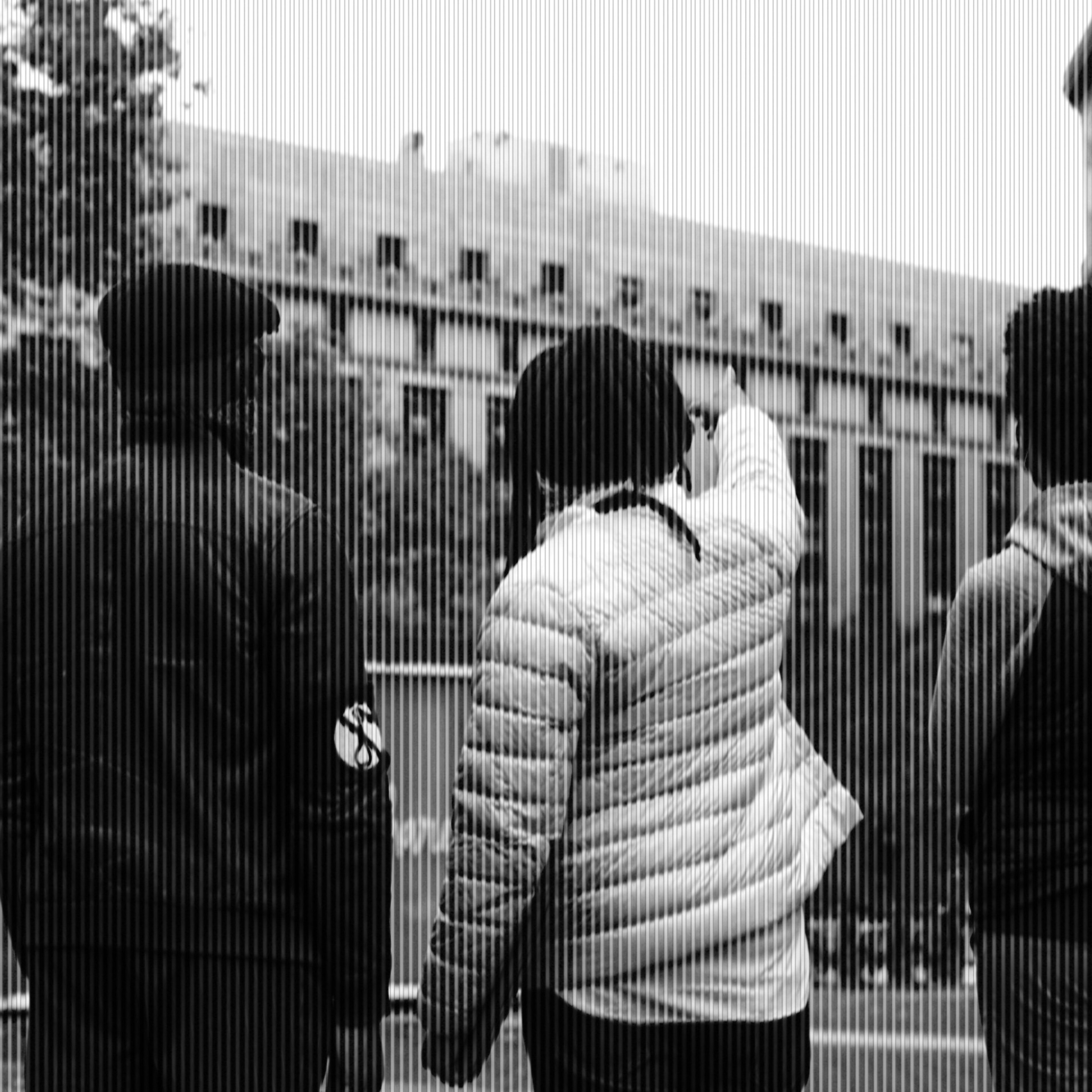 A black-and-white photo of three Black men seen from behind facing a building in the background. The one in the middle, artist Kamau Ware, points up at the building.