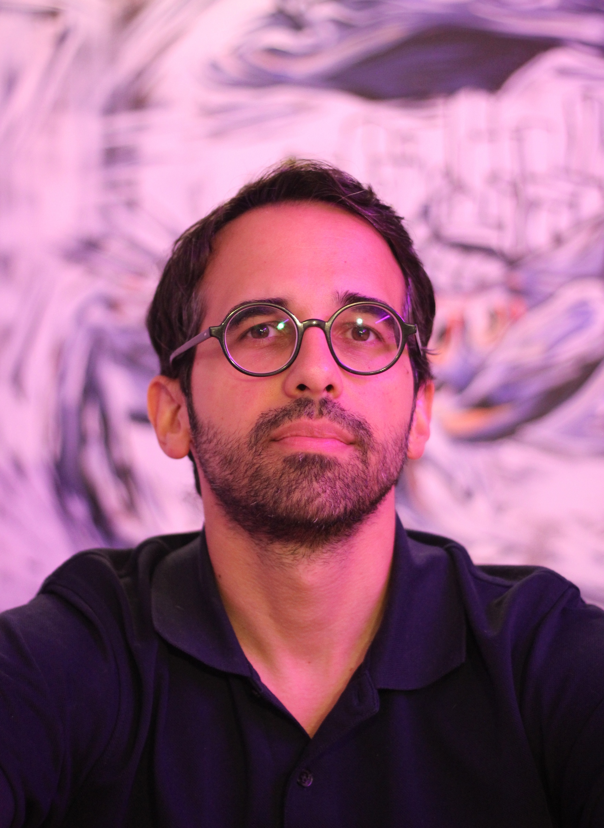 A photo of Oscar Oliver-Didier wearing glasses bathed in a pink light against a pink abstract background