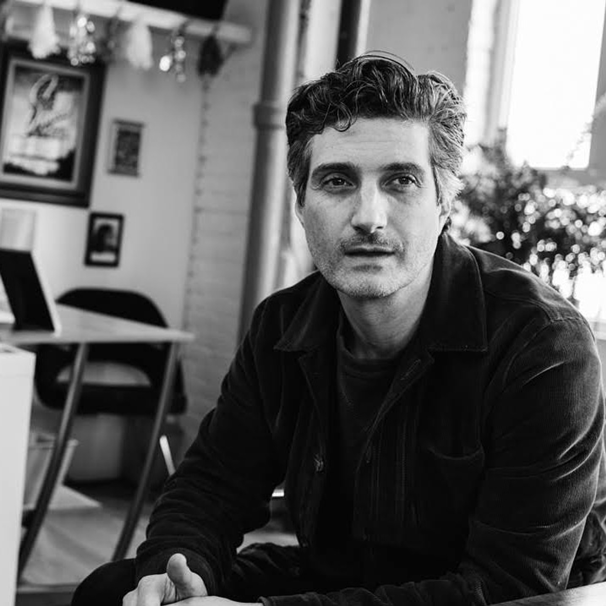 A black-and-white photo fo Noel Allain sitting with elbows on his knees, with a desk, chair, and plant in the background. 