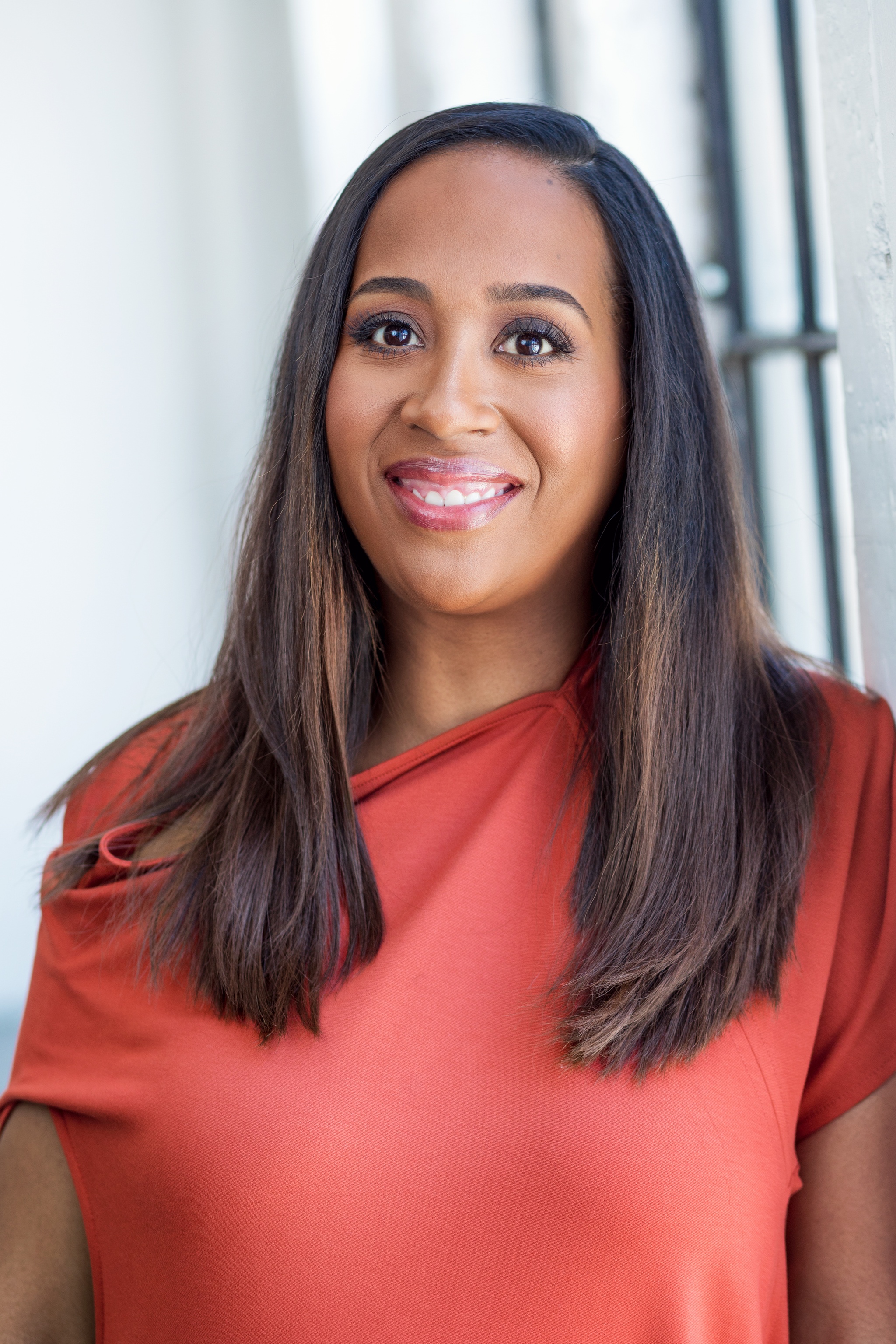 A photo of curator Naima J. Keith smiling at the camera and wearing a peach blouse with her hair falling over her shoulders. Natural light from a window fills the backhround.