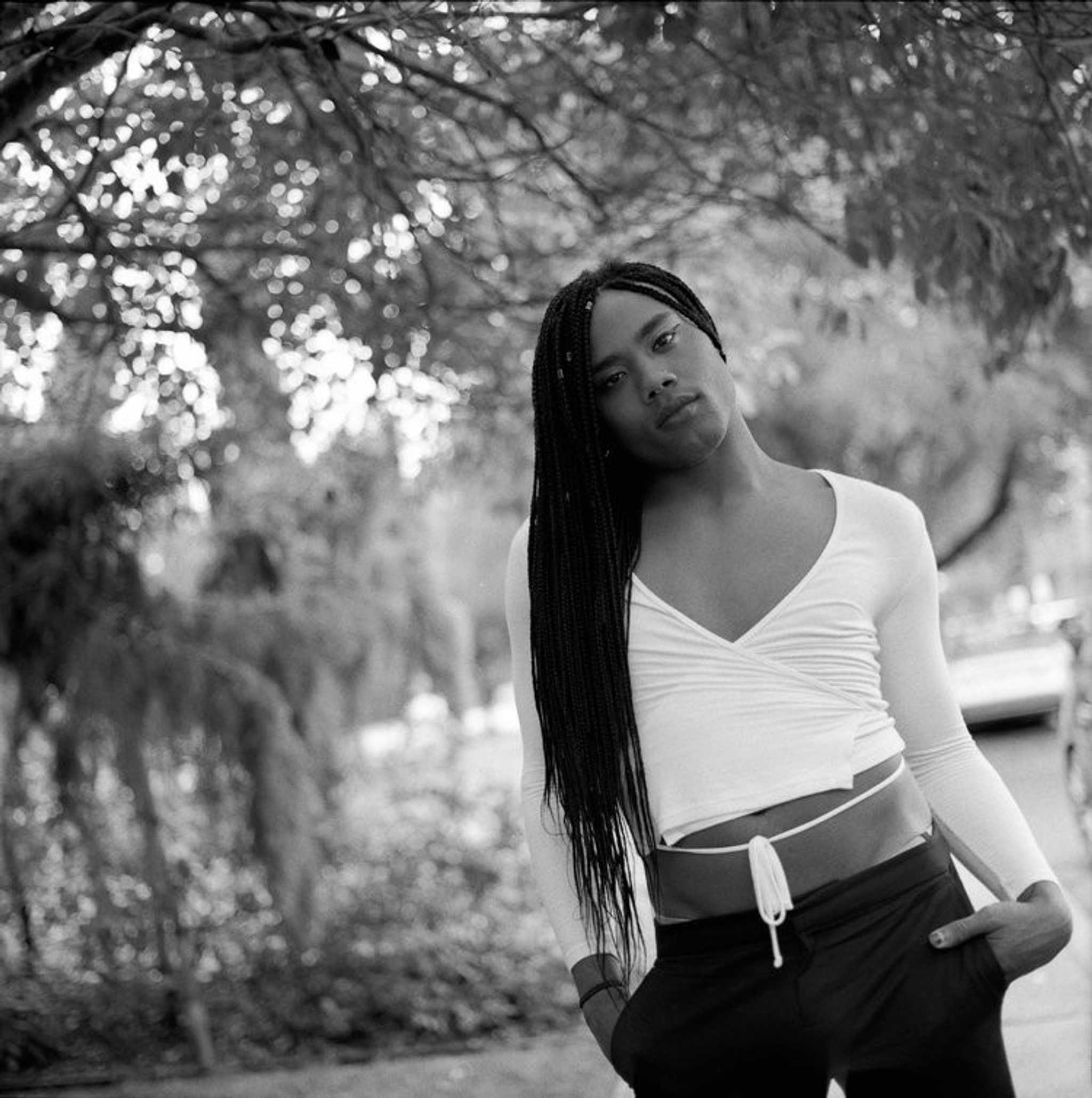 A portrait of Kiyan Williams standing against a background of trees with their hands casually in their pockets and head tilted to the side