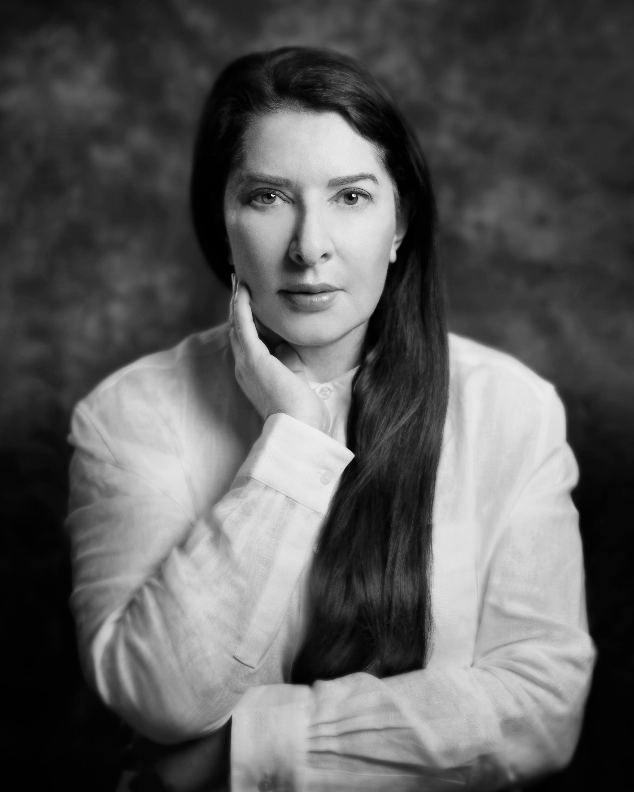 A portrait of Marina Abramovic. She sits facing us with one hand tucked under her chin supporting it. Her long dark hair falls over her left shoulder. She looks intently at us. 