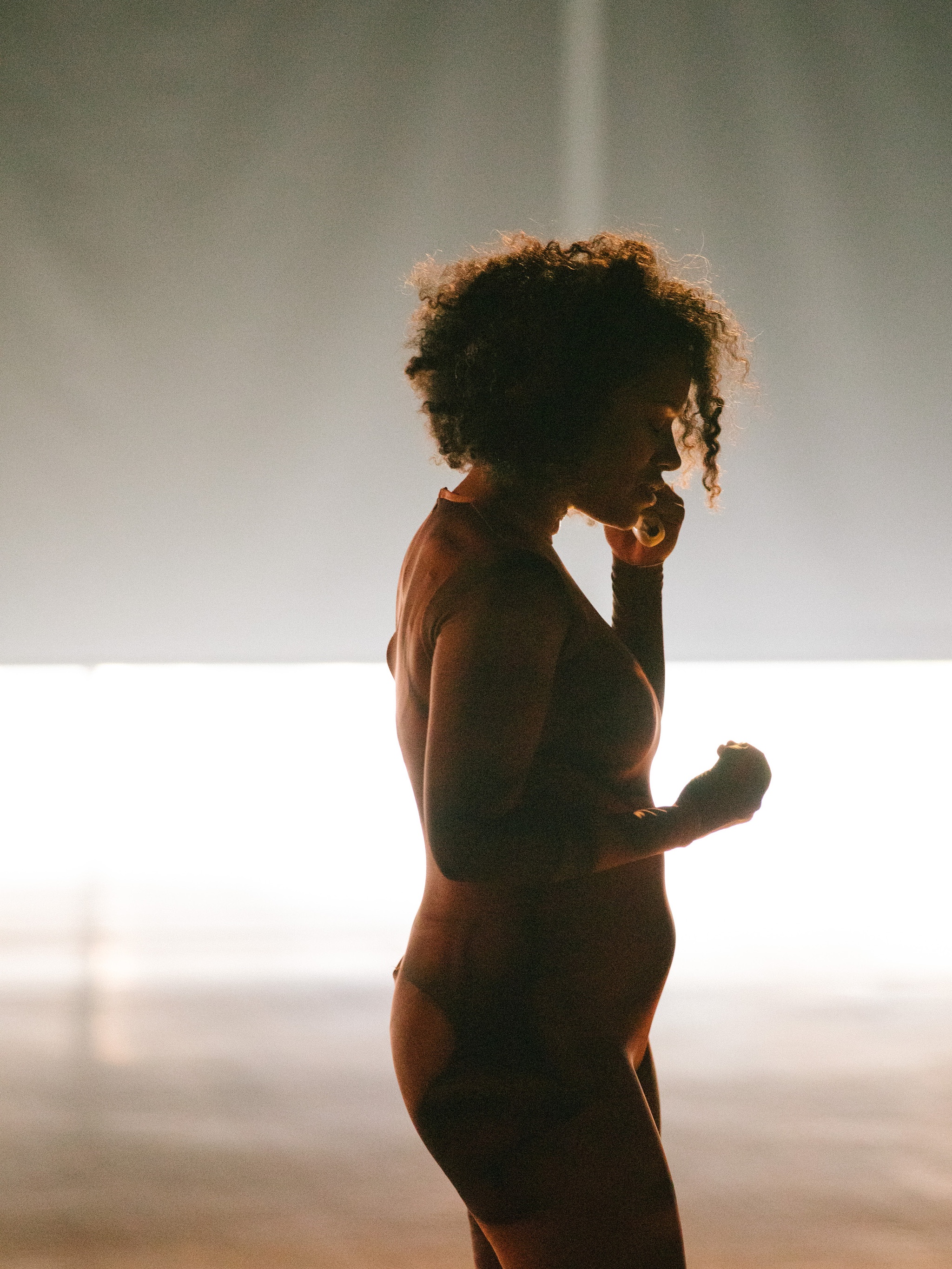 A dancer standing in profile silhouetted by light coming in through shaded windows behind her 