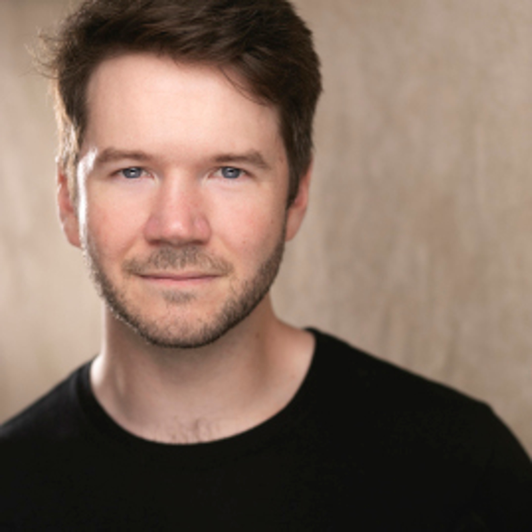 A portrait of artist James Caverly. Joey is a white man with brown hair parted to one side across his forehead. She smiles slightly and looks directly at us. He wears a black tshirt.