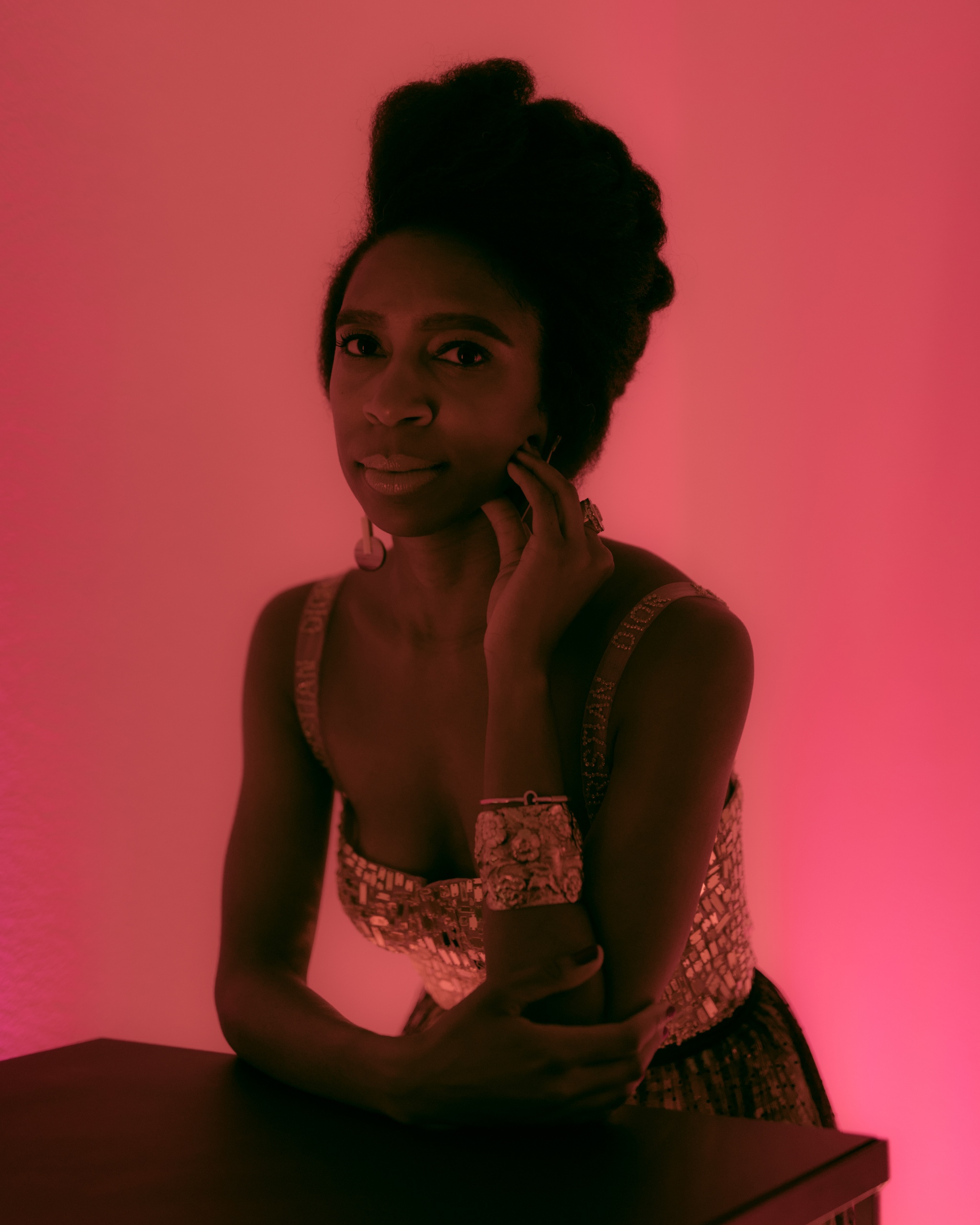 A portrait of DJ April Hunt backlit by a pink light. Hunt props her head on one hand and is wearing a gold sequined top.